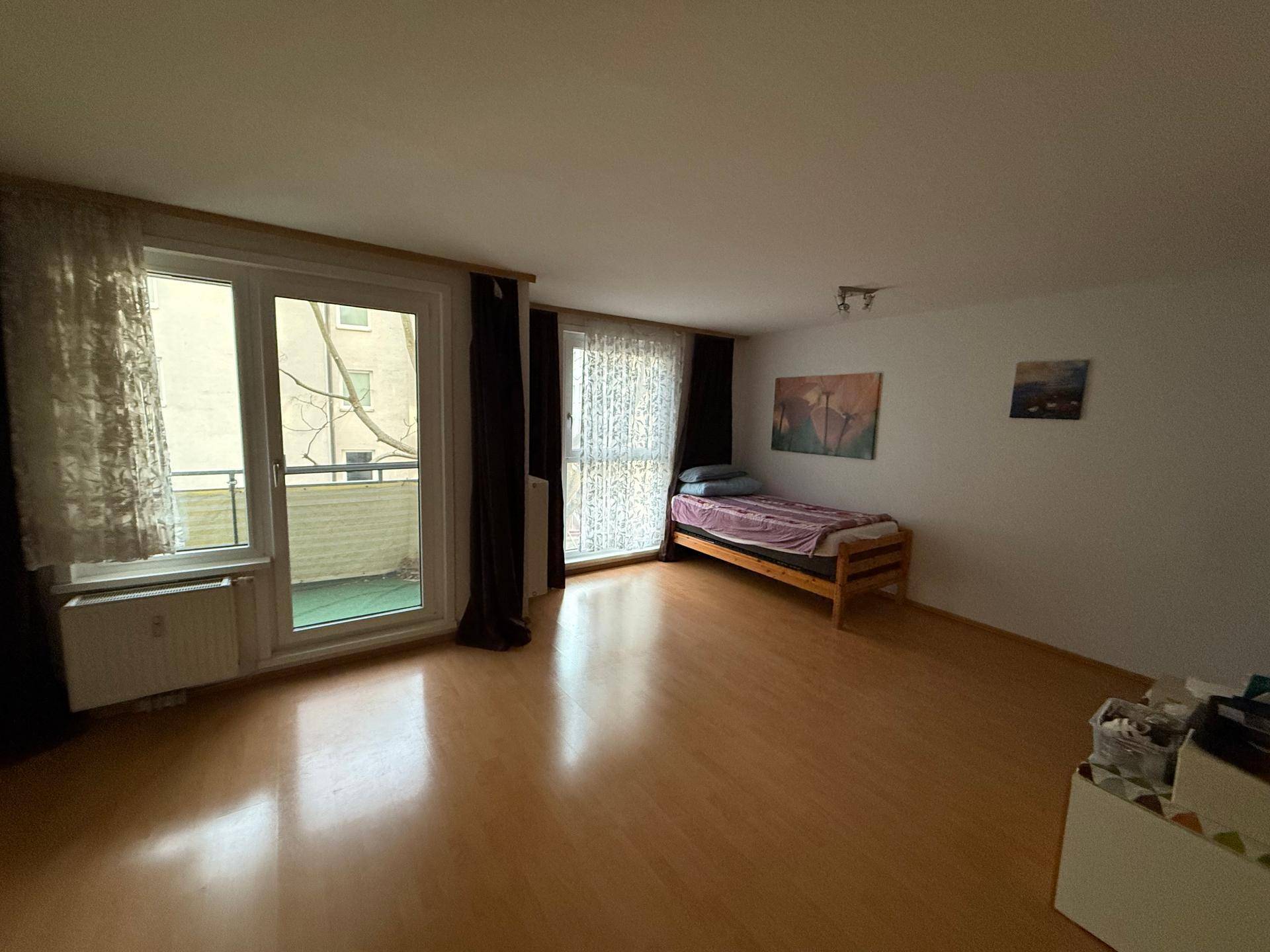 Geräumiger Wohnbereich mit Laminatboden, einem Bett und zwei großen Fenstern zum Balkon.