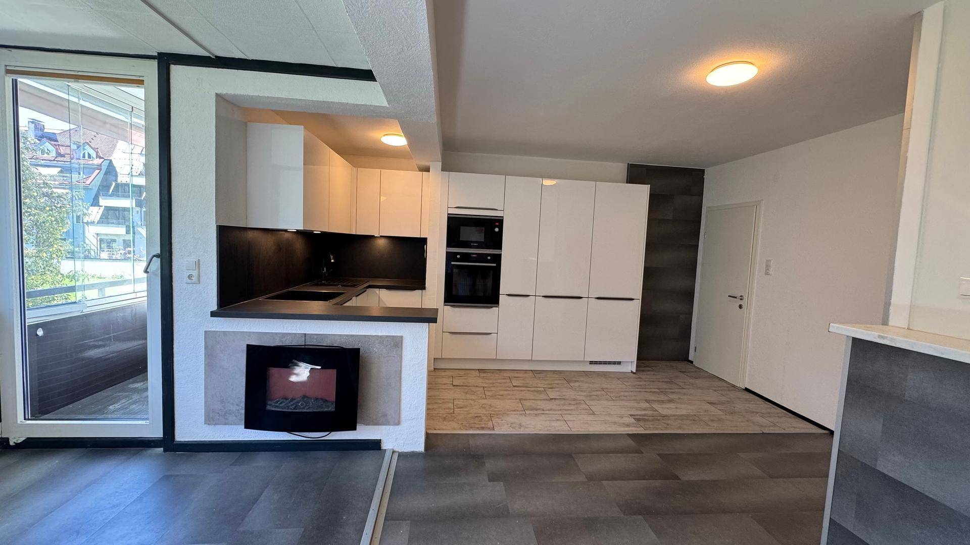 Offener Wohnbereich mit integrierter Küche, Kamin und Zugang zum verglasten Balkon.