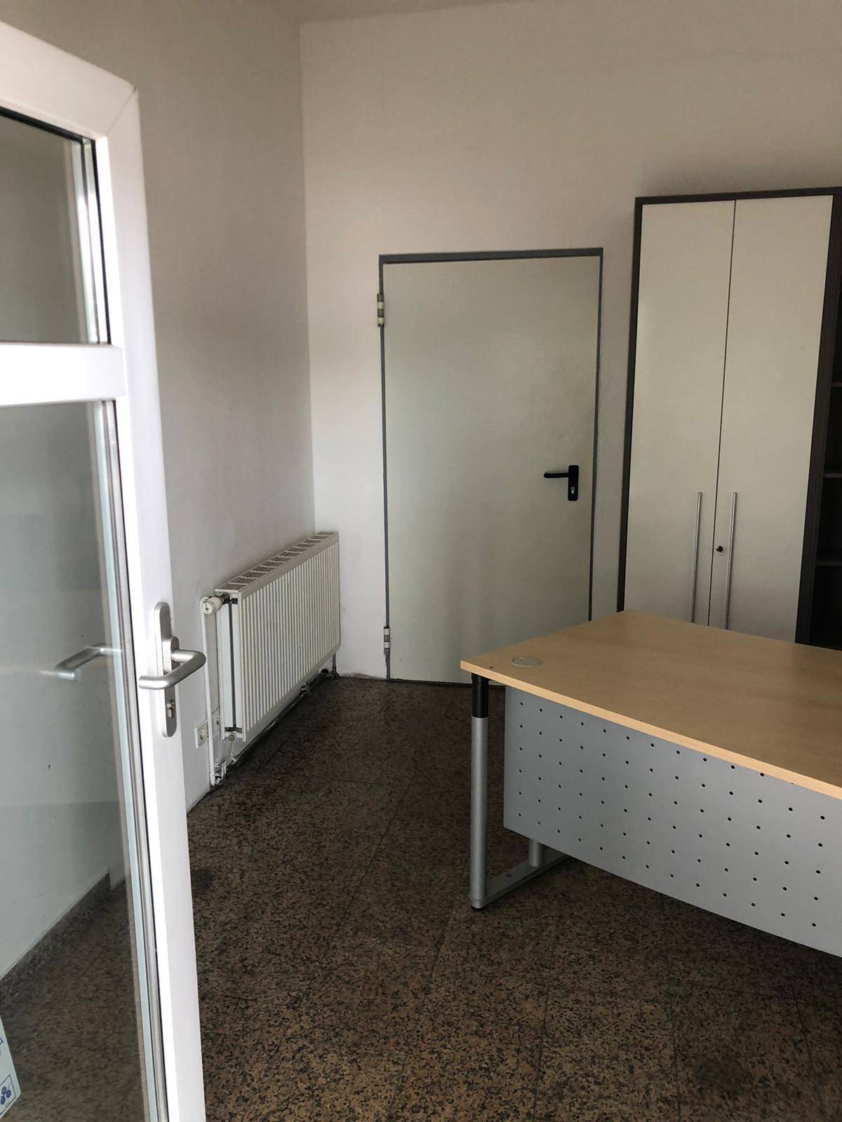 Büroraum mit Eingangstür, Heizkörper unter dem Fenster und funktionaler Büroeinrichtung an der Wand.