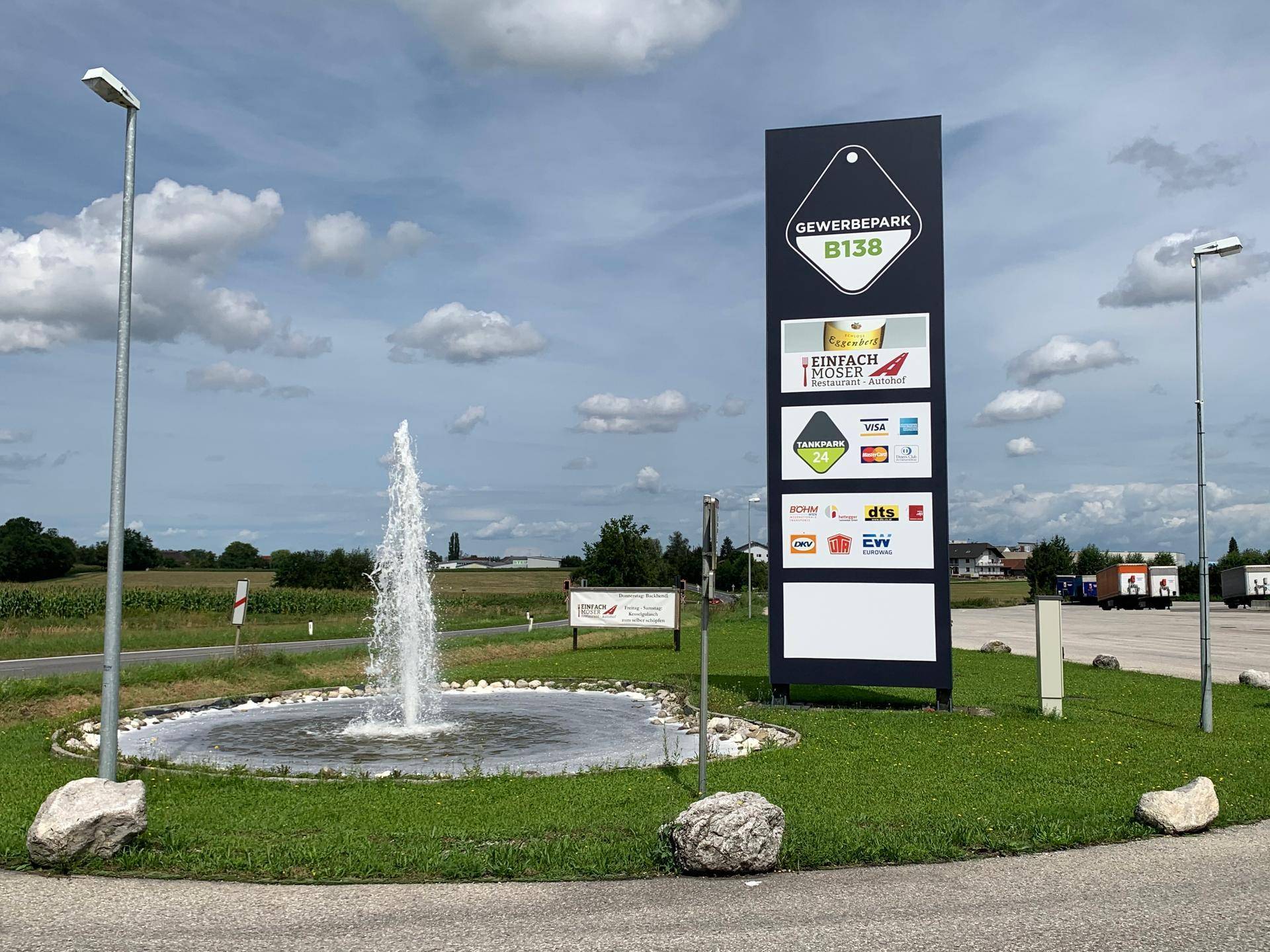 Großes Werbeschild des Gewerbeparks B138 mit Firmenlogos und einem dekorativen Springbrunnen in einer Grünanlage.