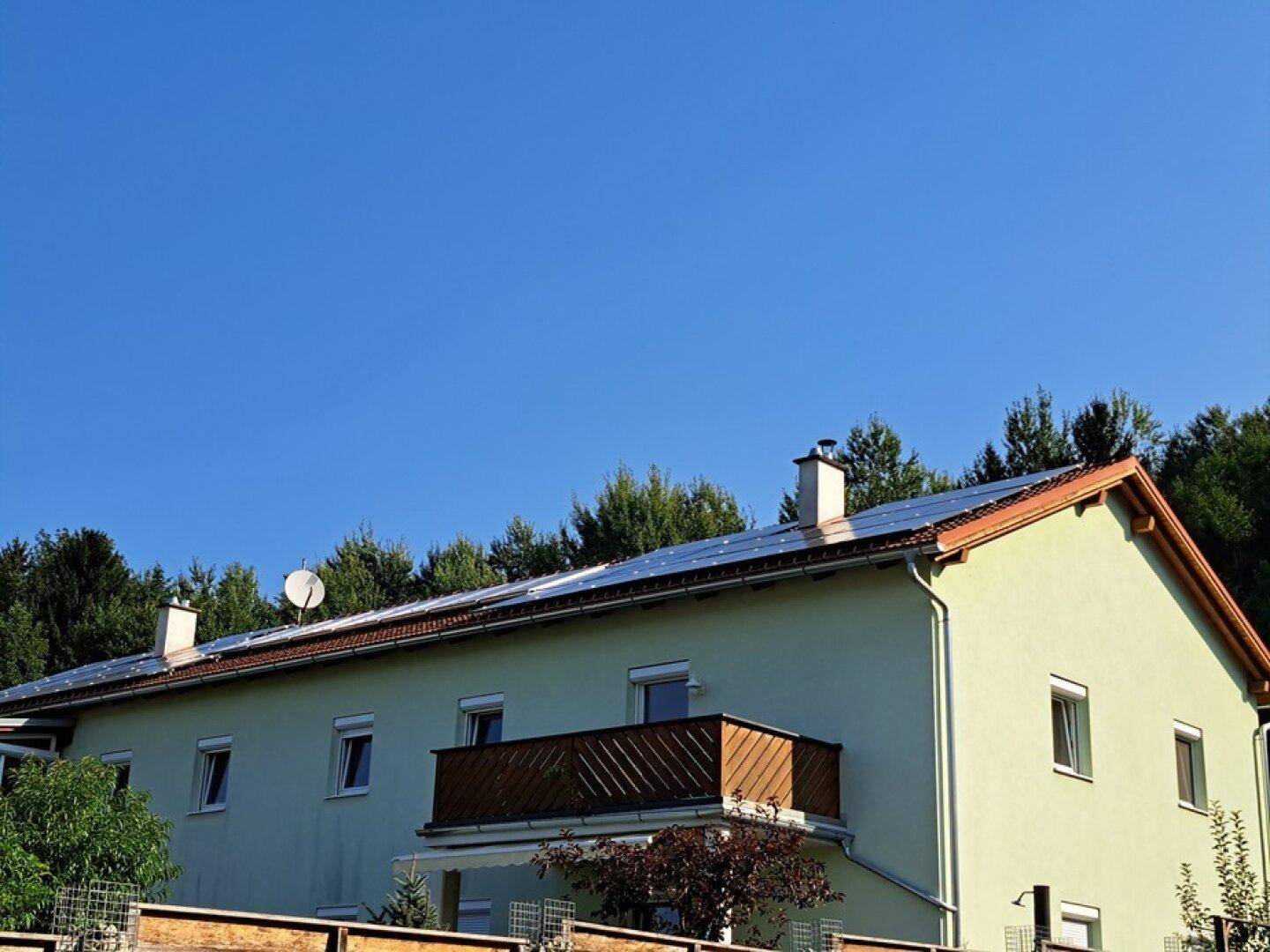 Hausfassade mit Balkon und Photovoltaikanlage auf dem Satteldach unter blauem Himmel.