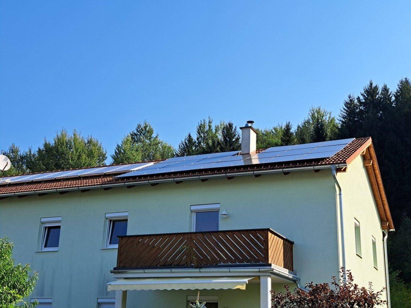 Hausfassade mit Balkon und Photovoltaikanlage auf dem Satteldach unter blauem Himmel.