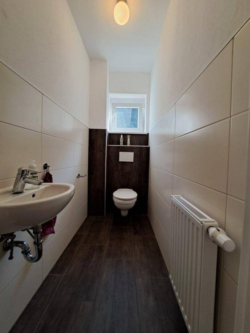 Schmales Gäste-WC mit wandhängender Toilette, kleinem Waschbecken und Fenster.