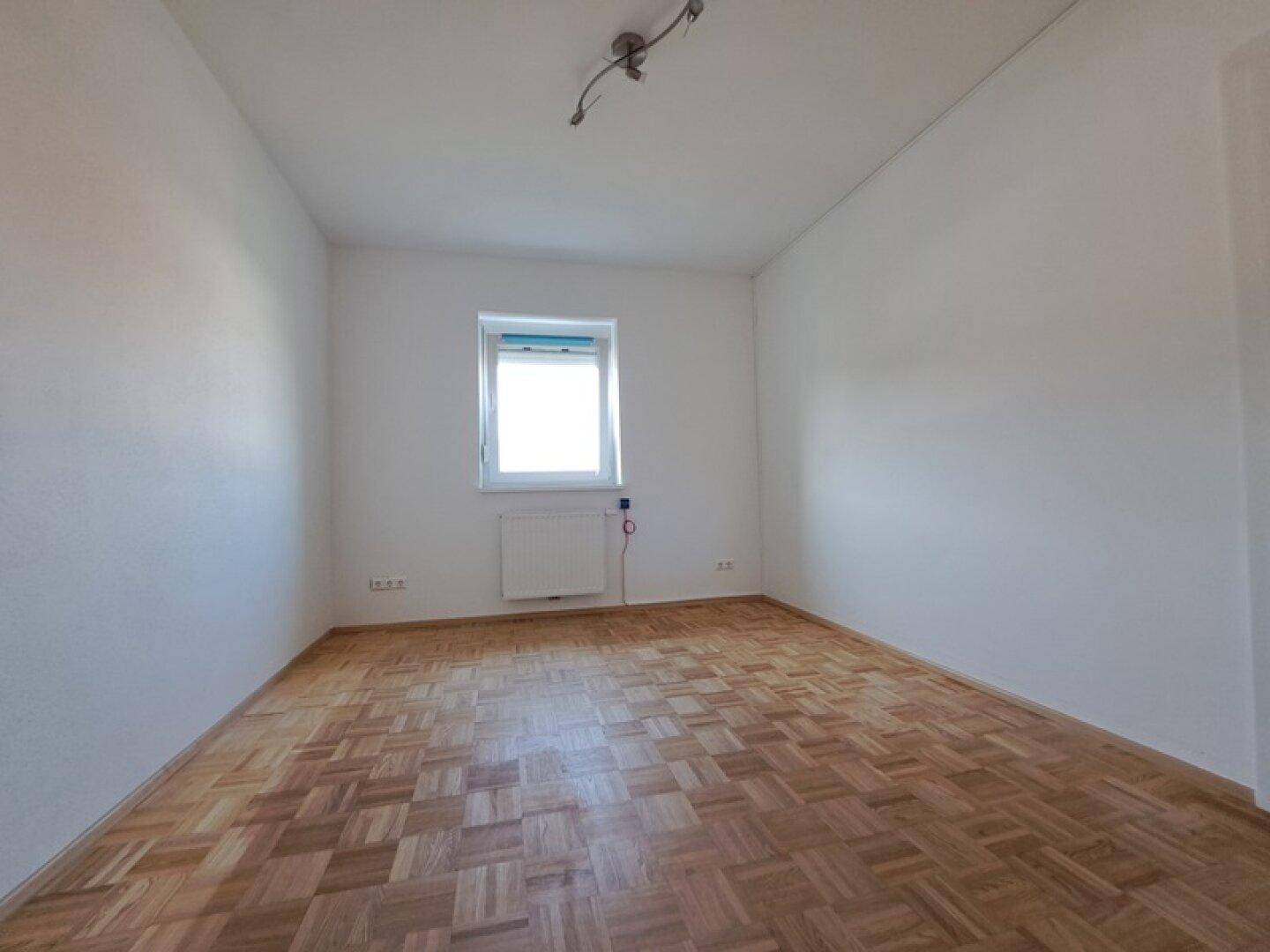 Helles Zimmer mit Parkettboden und einem Fenster, das viel Tageslicht hereinlässt.