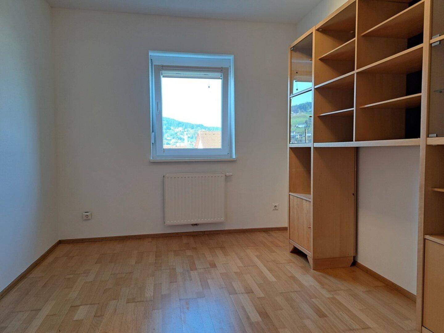 Geräumiges Zimmer mit Parkettboden, Fenster und einem großen, hellen Holzregal.