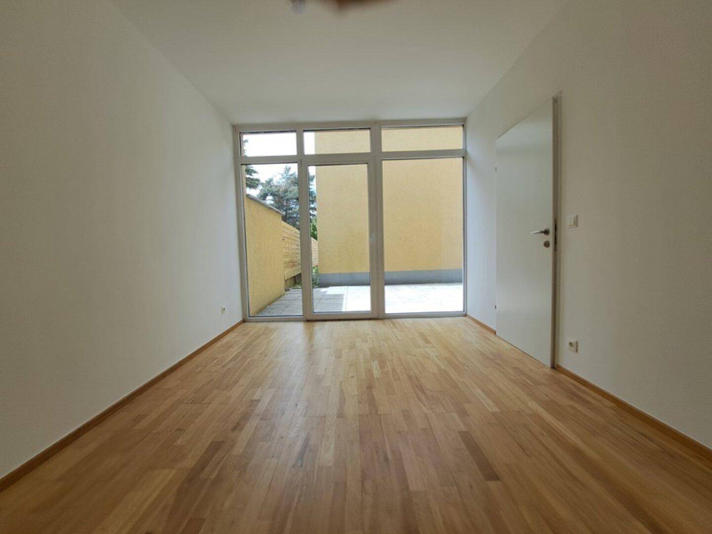 Heller Raum mit Holzboden und großer Fensterfront, die Zugang zu einer Terrasse bietet.