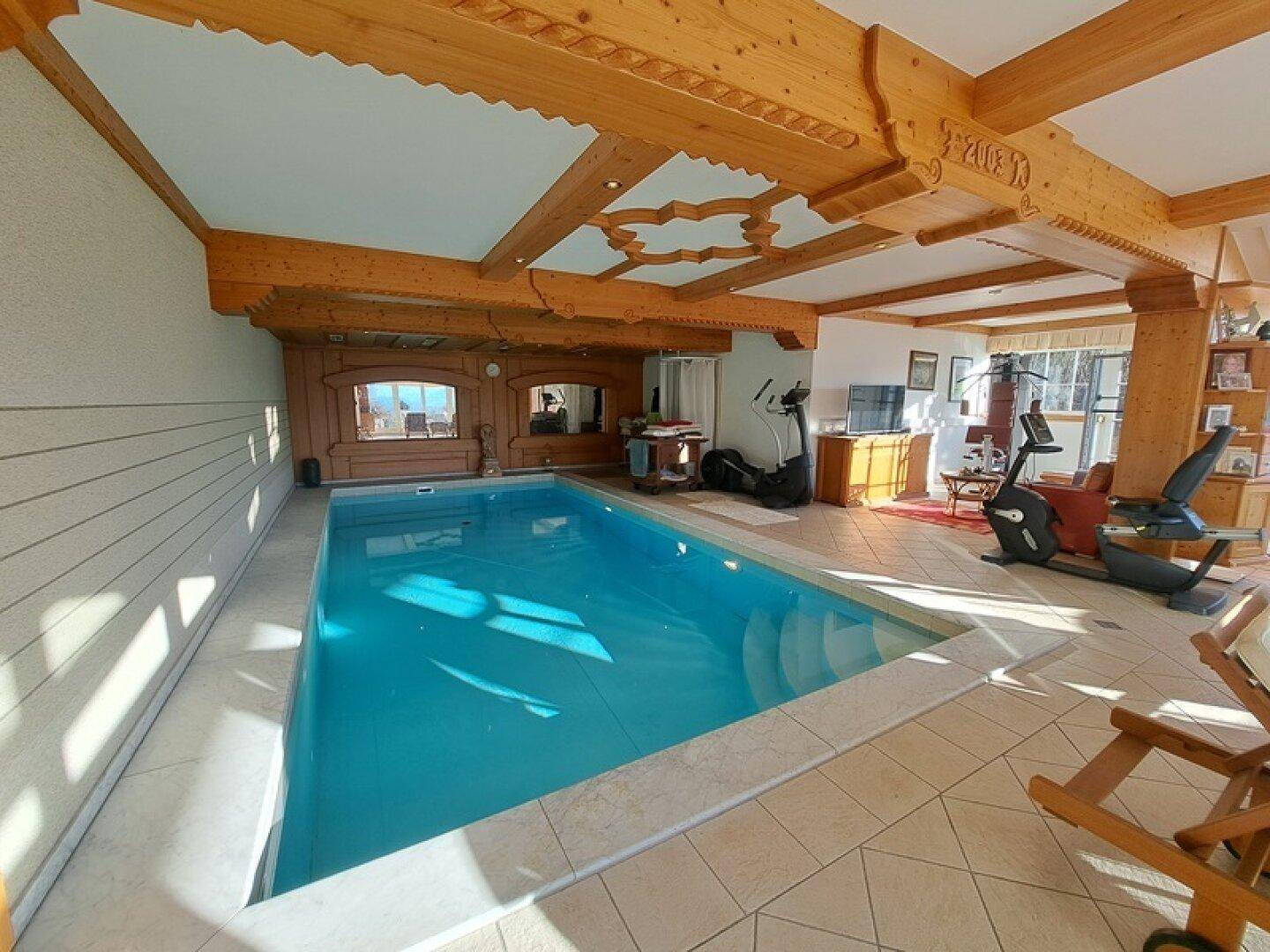 Innenpoolbereich mit Fitnessgeräten, Holzbalkendecke und großen Fenstern.