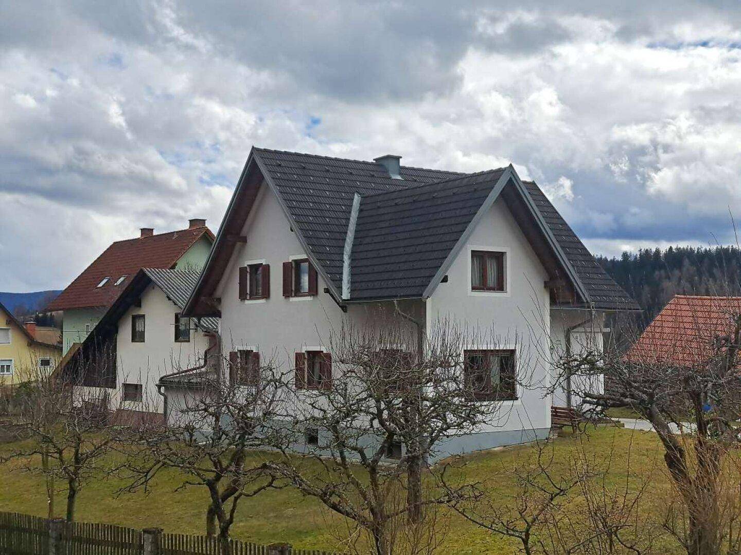 Das Haus in ländlicher Umgebung, umgeben von Bäumen und einem Holzzaun unter bewölktem Himmel.