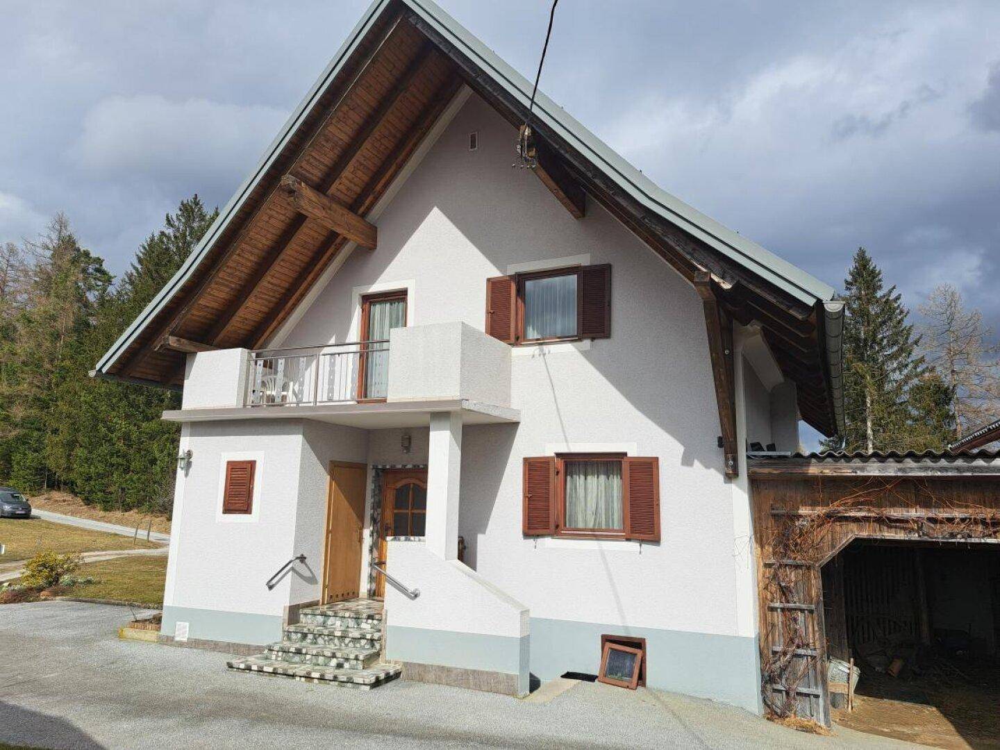 Vorderansicht des Hauses mit Balkon, Holzdetails und traditionellen Fensterläden vor bewölktem Himmel.