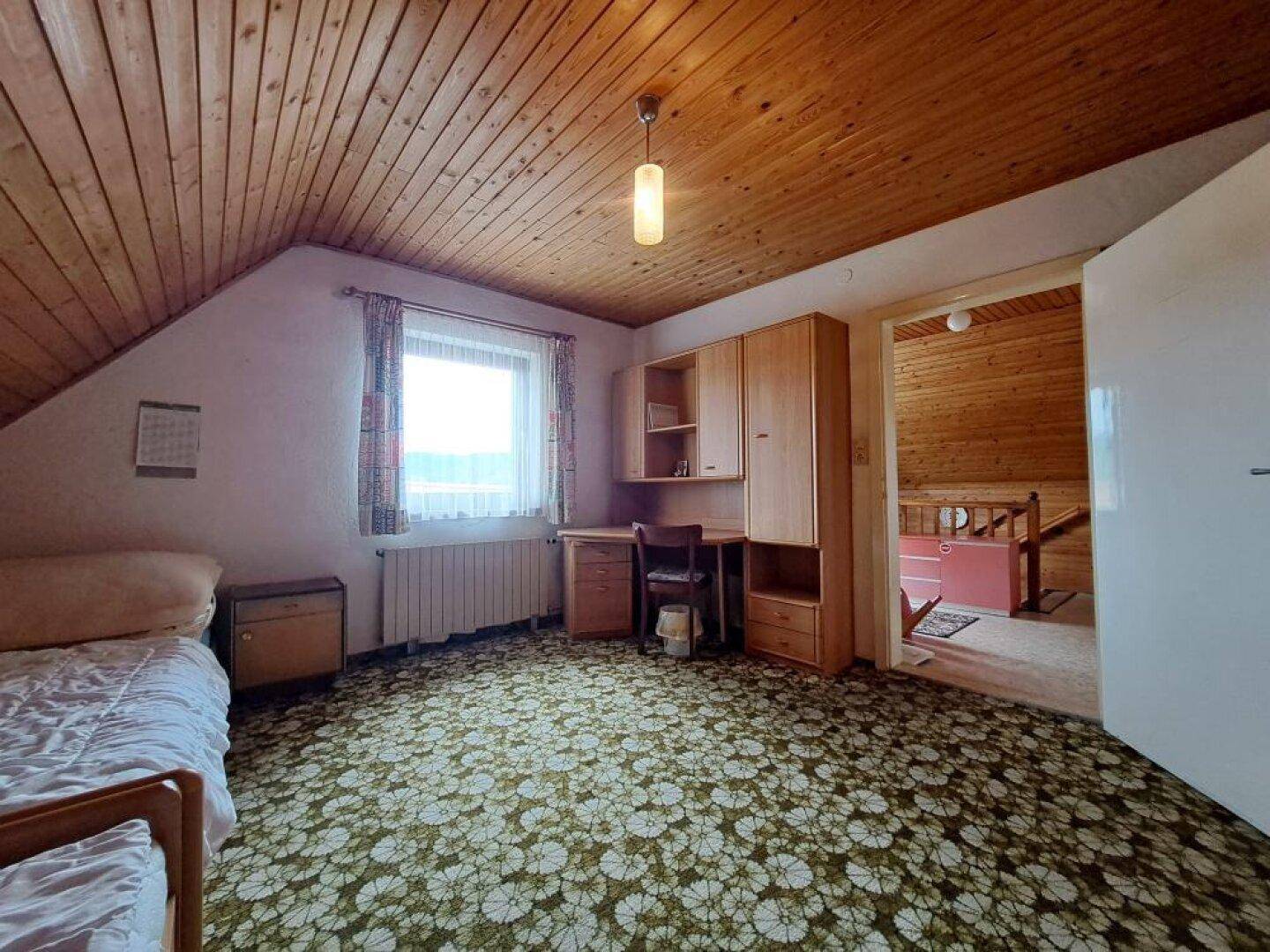 Schlafzimmer mit Holzdecke, gemustertem Teppich und Schreibtisch unter dem Fenster.