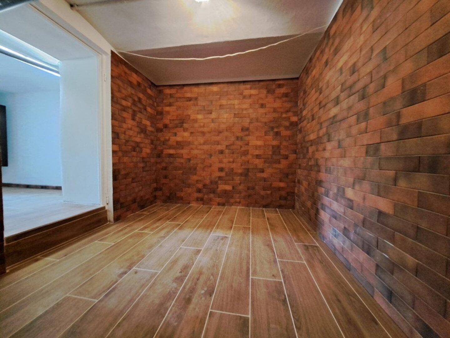 Schmaler Innenraum mit dekorativen Ziegelwänden und Bodenfliesen in Holzoptik, der zu einem weiteren Zimmer führt.