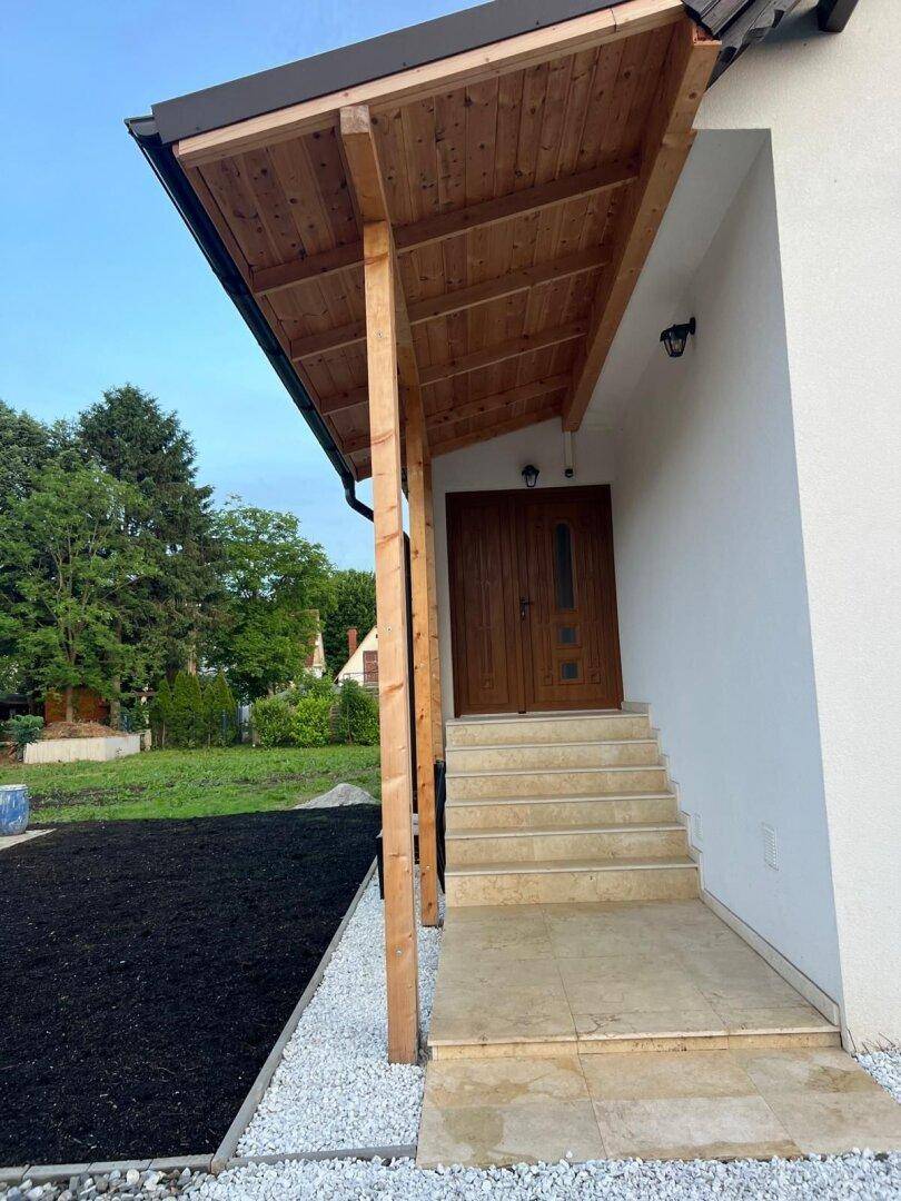 Überdachter Hauseingang mit Holzstützen und Steintreppe, umgeben von Kies und Grünflächen.
