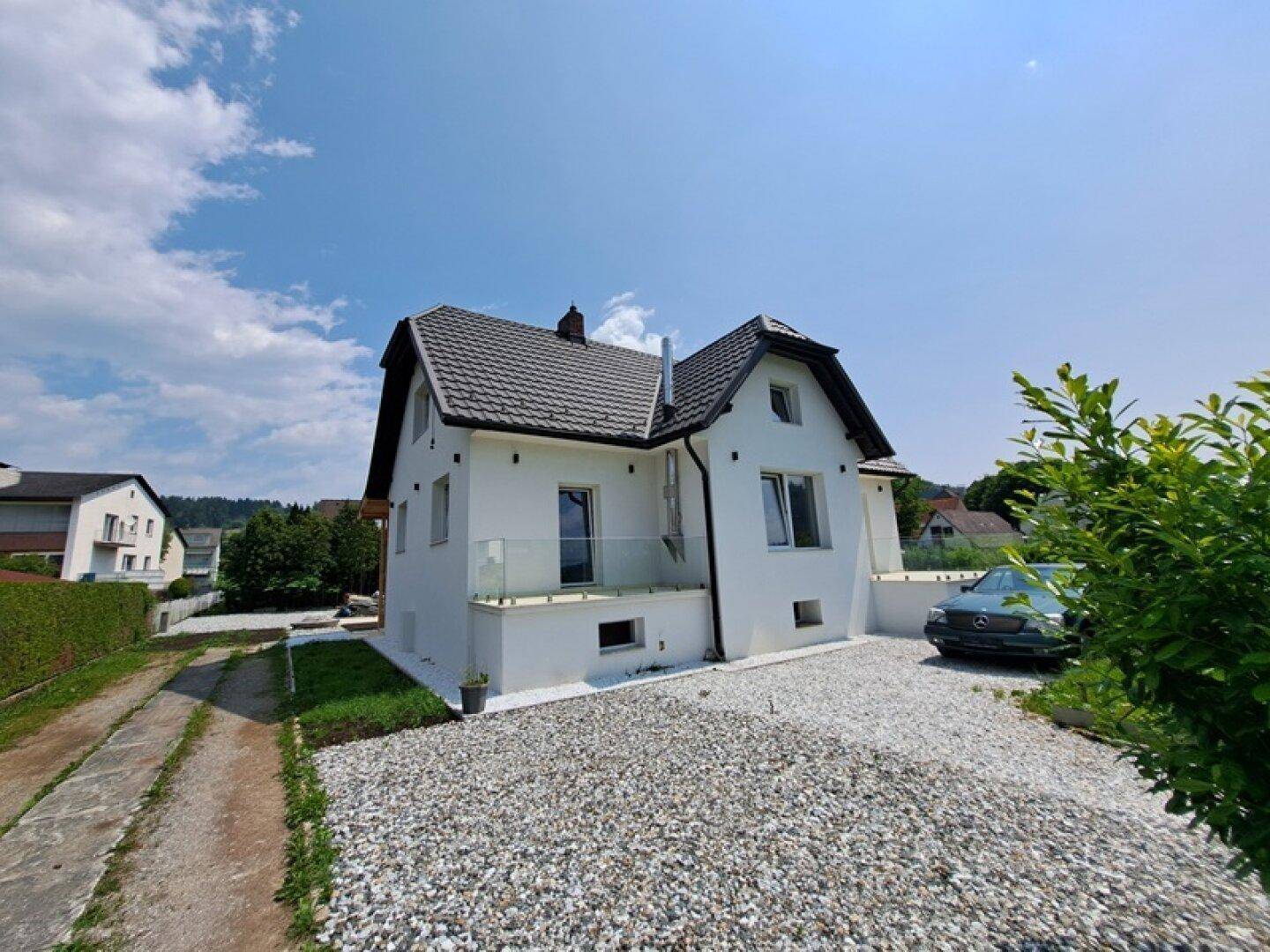 Gesamtansicht des Hauses mit Kiesauffahrt und umliegender grüner Landschaft unter blauem Himmel.