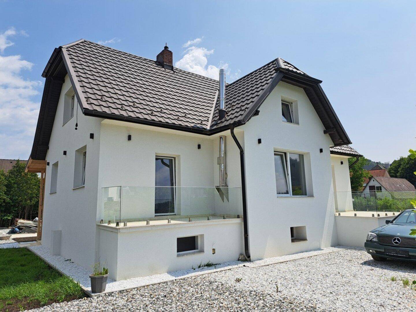 Seitenansicht des Hauses mit moderner Glasbrüstung und Kiesauffahrt unter strahlendem Himmel.