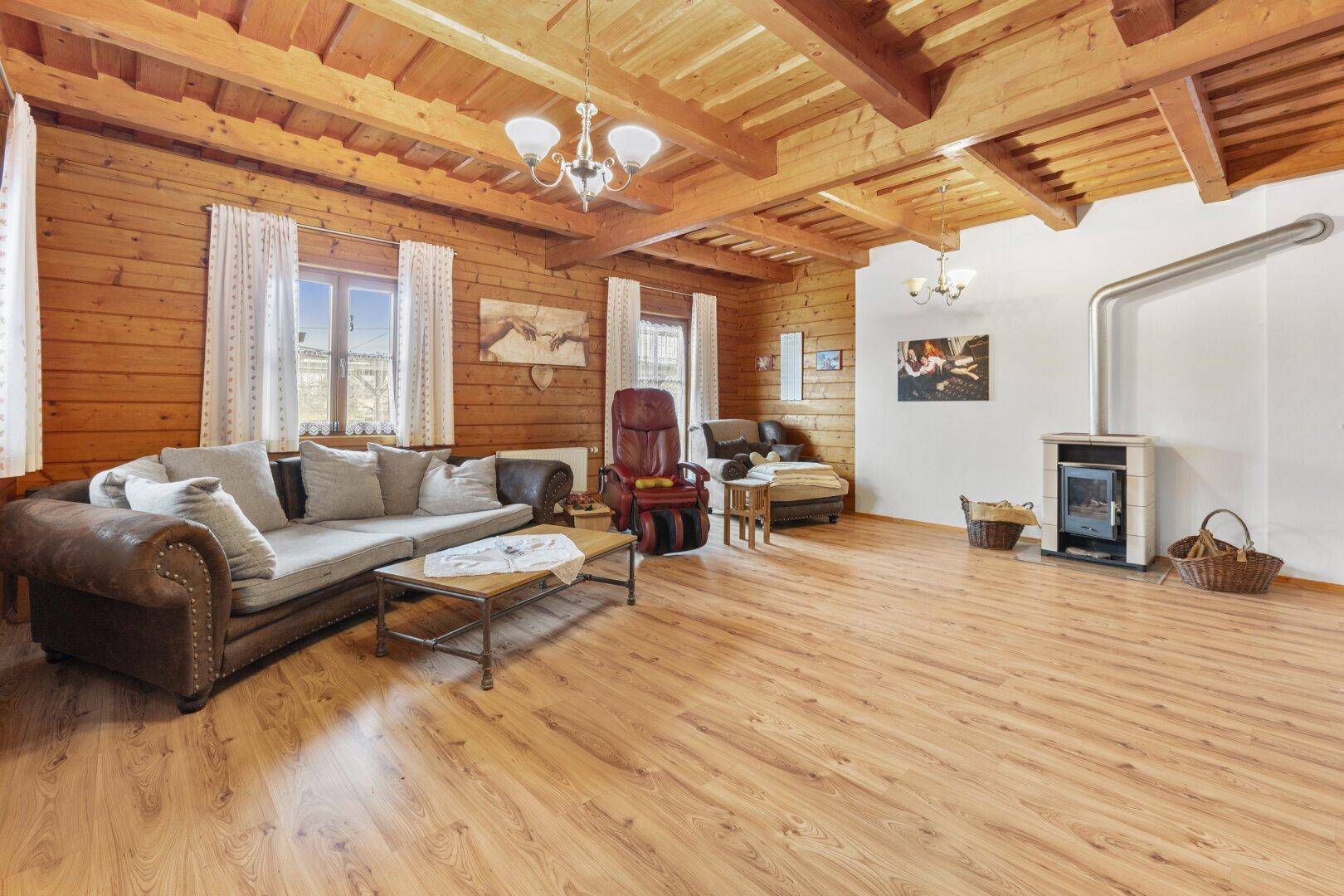 Helles Wohnzimmer mit Holzwänden, Holzbalkendecke, Laminatboden und einem modernen Kaminofen.