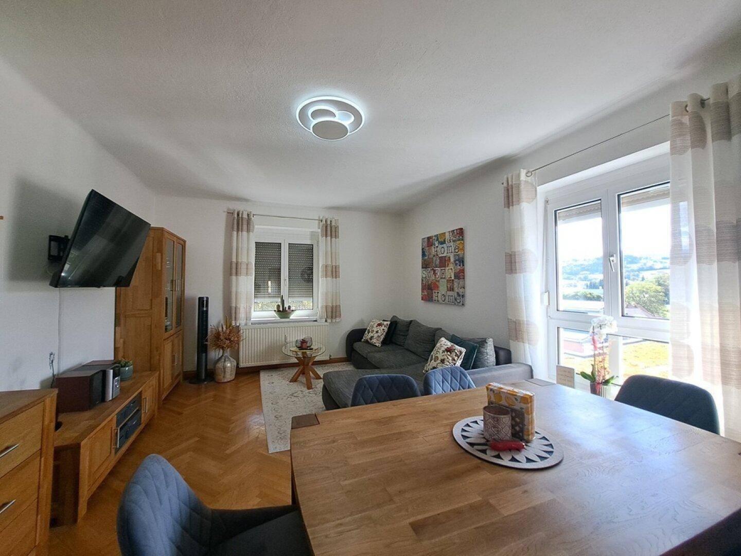 Gemütlicher Wohn-Essbereich mit Holztisch, grauem Sofa und Parkettboden, hell durch große Fenster mit grüner Aussicht.