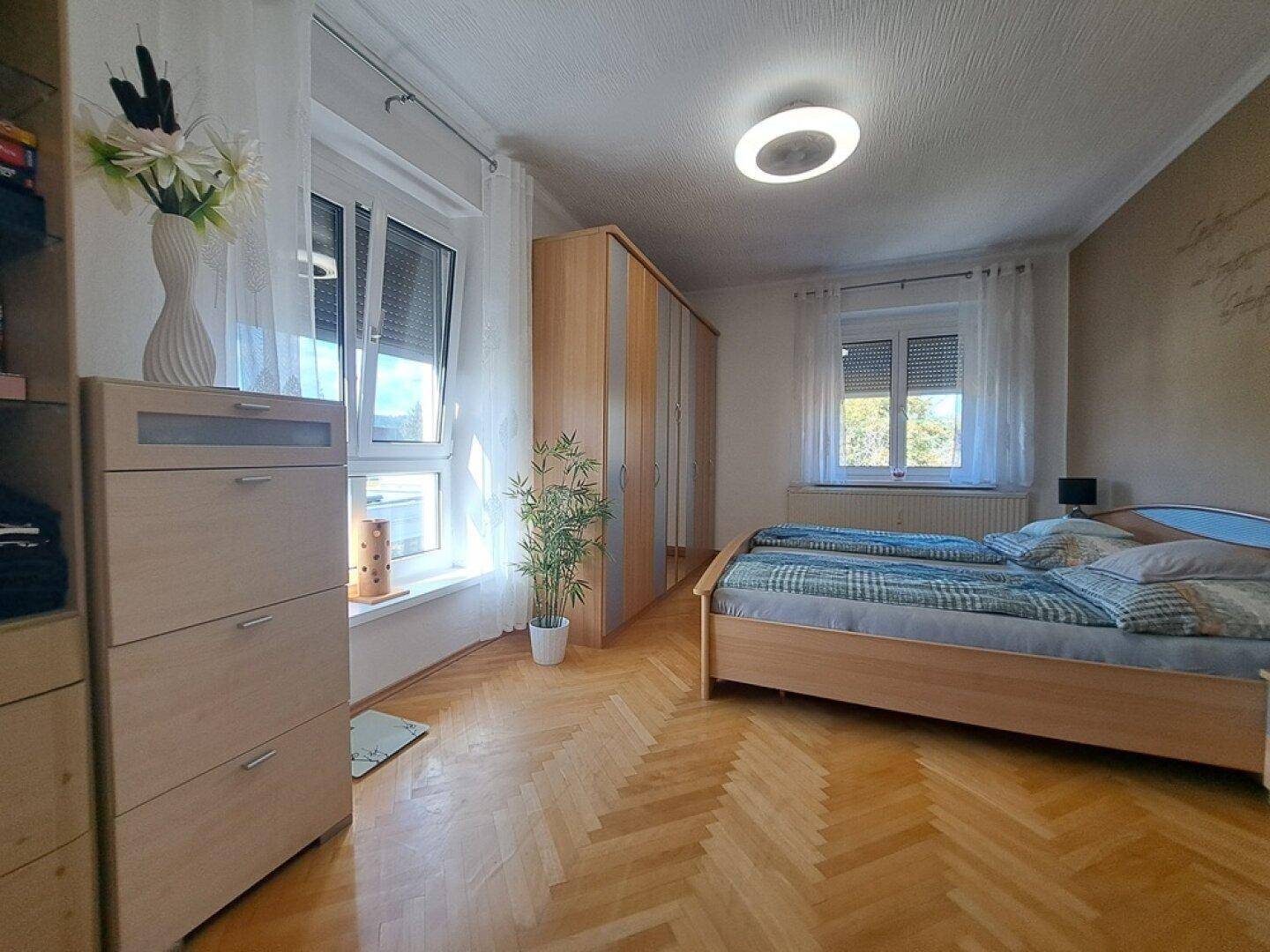 Helles Schlafzimmer mit Doppelbett, Holzschränken und Parkettboden, durch zwei Fenster gut belichtet.