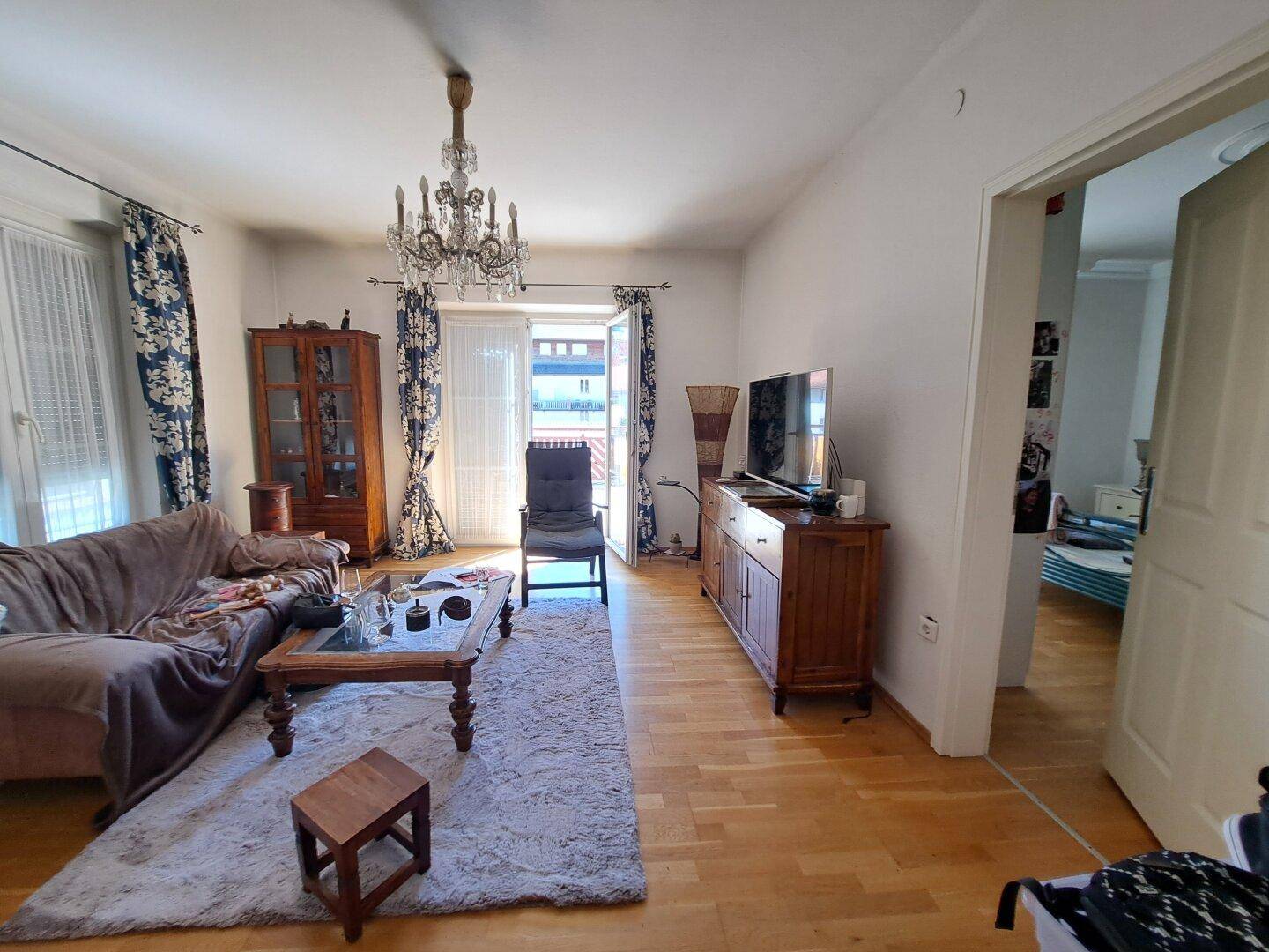 Helles Wohnzimmer mit Holzboden, Sofa, Kronleuchter und Balkonzugang.