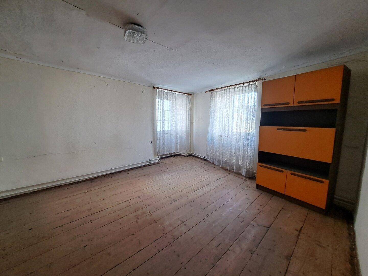 Leeres Zimmer mit Holzdielenboden, zwei Fenstern und einem einfachen Schrank, der Renovierung bedarf.