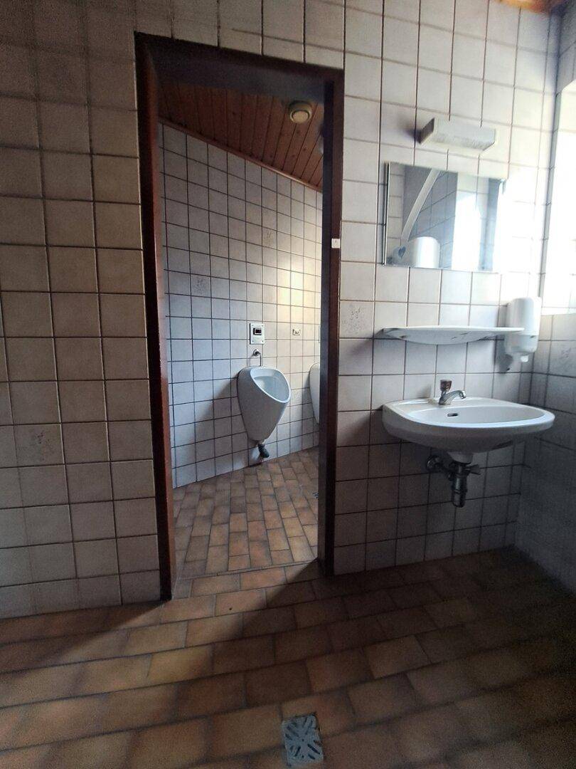 Einfache öffentliche Toilette mit Fliesenboden, Urinal und Waschbecken, in funktionalem Zustand.