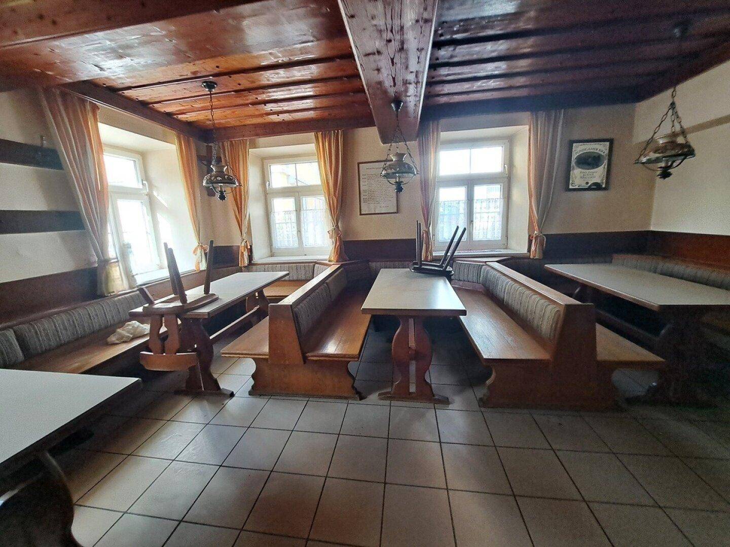 Gemütlicher Gastraum mit Holzbänken, Tischen und großen Fenstern, die viel Tageslicht hereinlassen.
