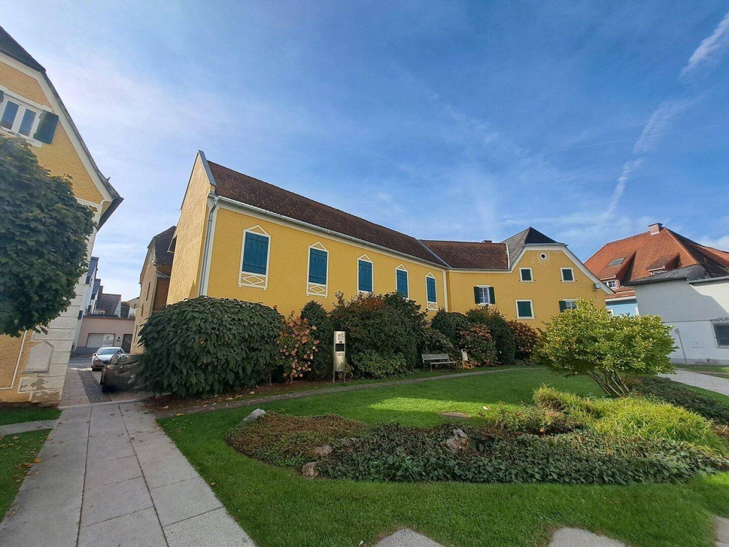Seitenansicht eines historischen gelben Gebäudes mit grünen Fensterläden und einem grünen Gartenbereich.