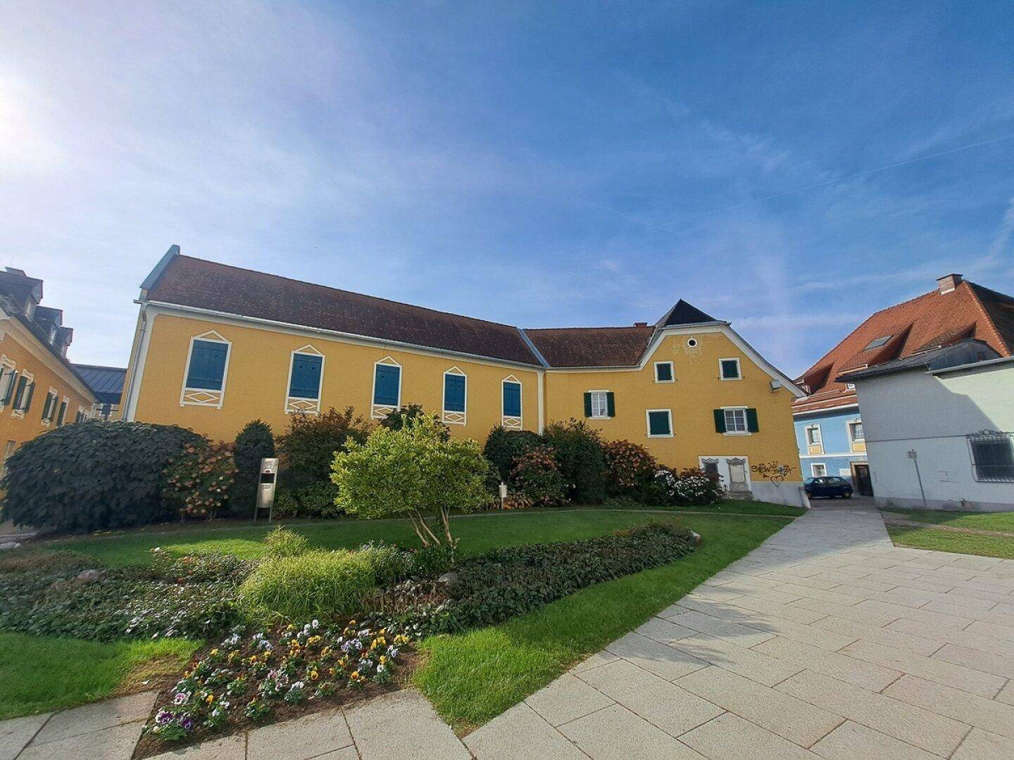 Historisches gelbes Gebäude mit grünen Fensterläden, umgeben von einem gepflegten Gartenbereich unter blauem Himmel.