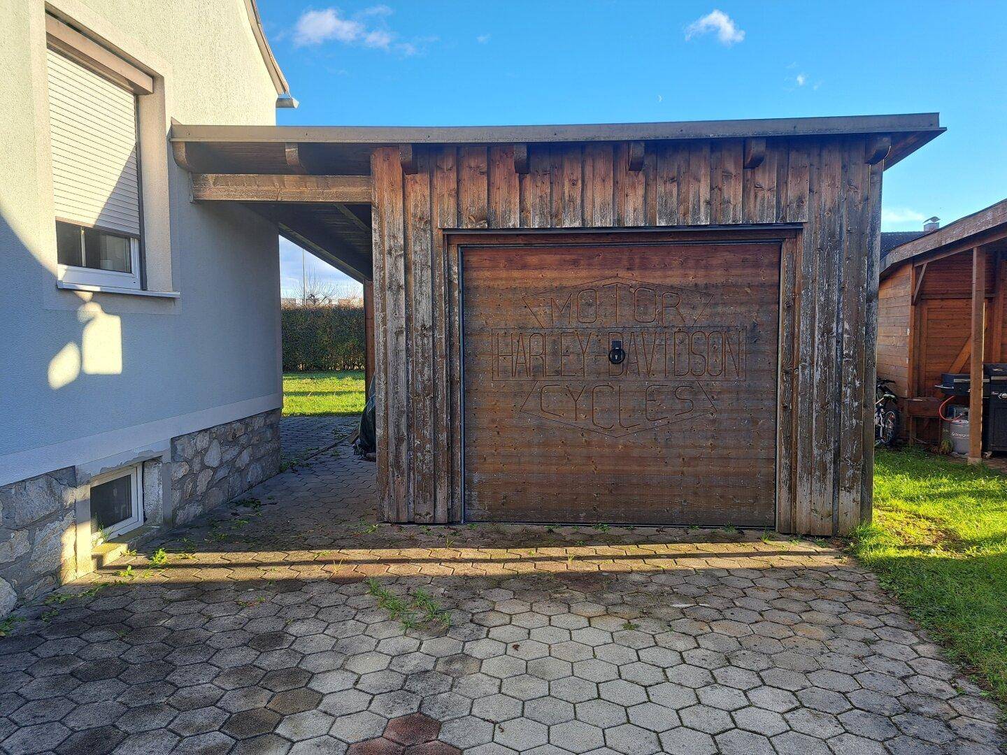 Hölzerne Garage mit Carport und gepflasterter Einfahrt neben dem Haus.