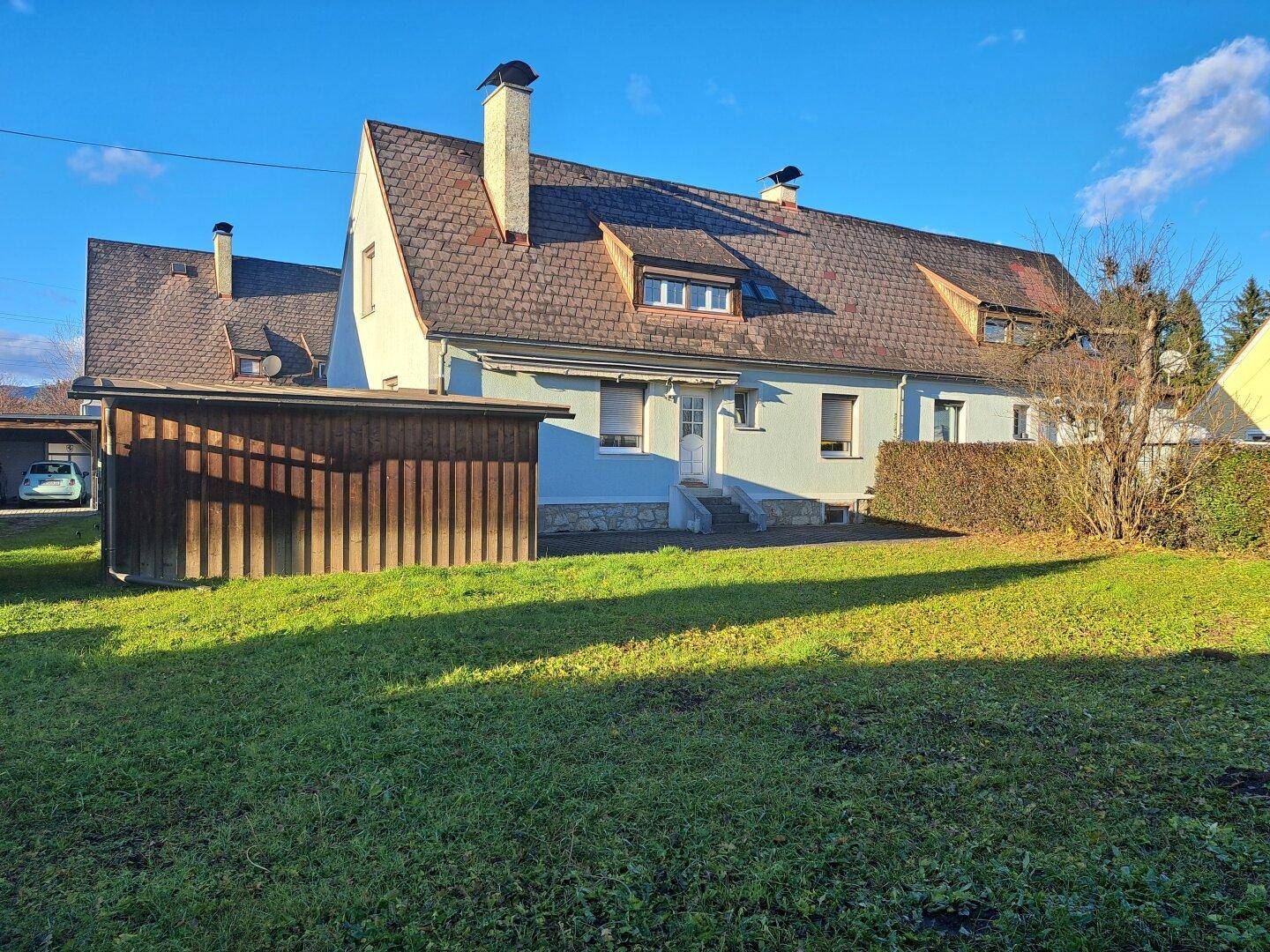 Hausfassade mit Garten, Rasenfläche und Holzschuppen unter blauem Himmel.