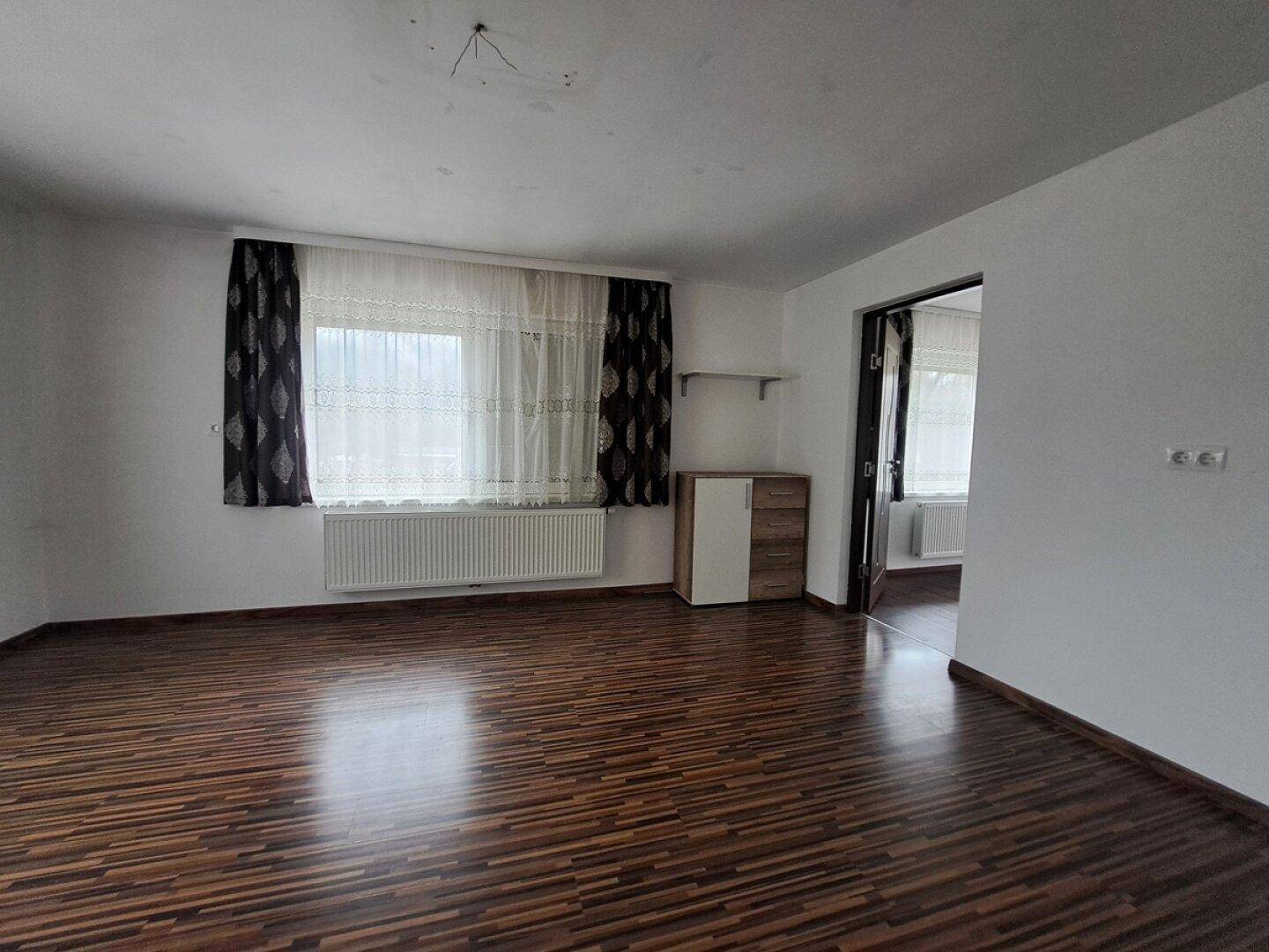 Geräumiges Zimmer mit dunklem Laminatboden, großen Fenstern und Blick in einen weiteren Raum.