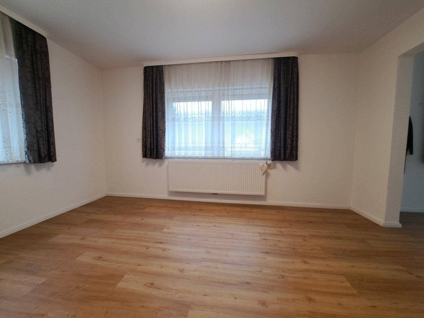 Leeres Zimmer mit hellem Laminatboden, großen Fenstern und einem Heizkörper unter dem Fenster.