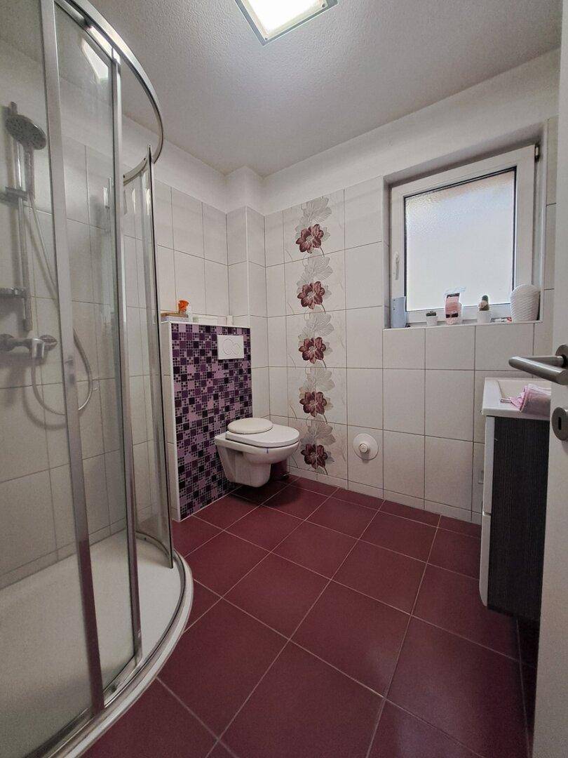 Badezimmer mit Duschkabine, weißen und violetten Fliesen sowie einem Fenster für Tageslicht.