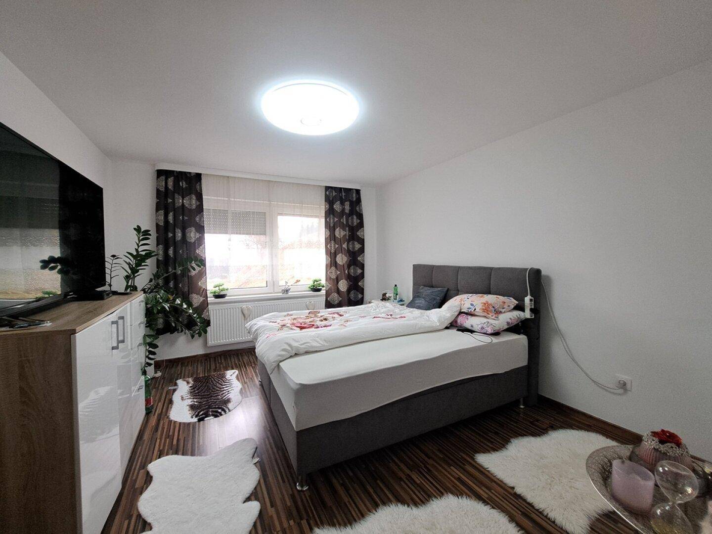 Schlafzimmer mit Doppelbett, dunklem Laminatboden und hellem Fenster für natürliche Beleuchtung.