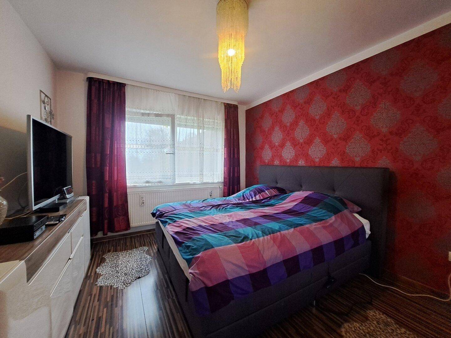 Schlafzimmer mit Doppelbett, dunklem Laminatboden und einer auffälligen roten Mustertapete.