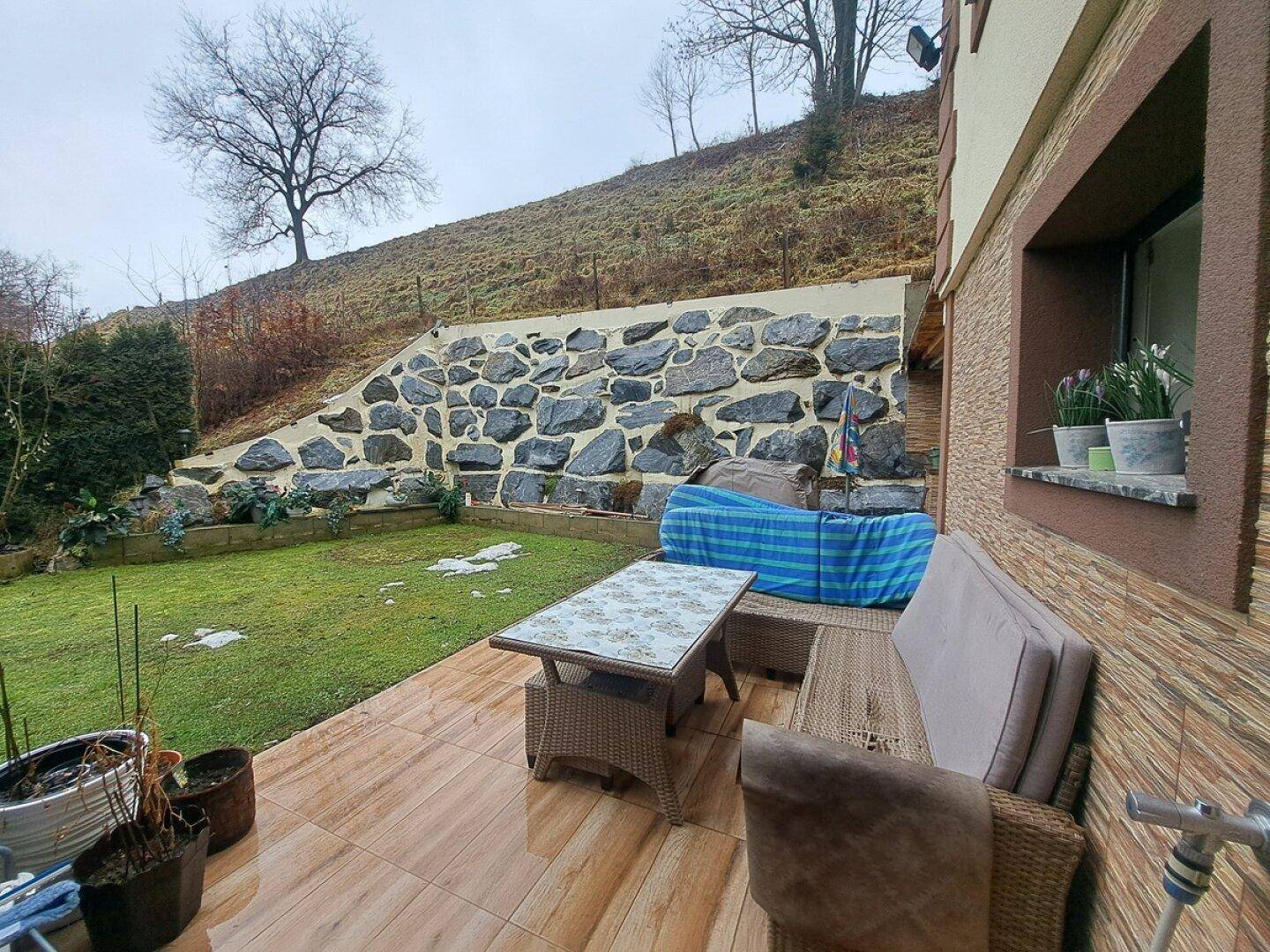 Terrasse mit Gartenmöbeln und Blick auf eine dekorative Steinmauer und den grünen Rasen.