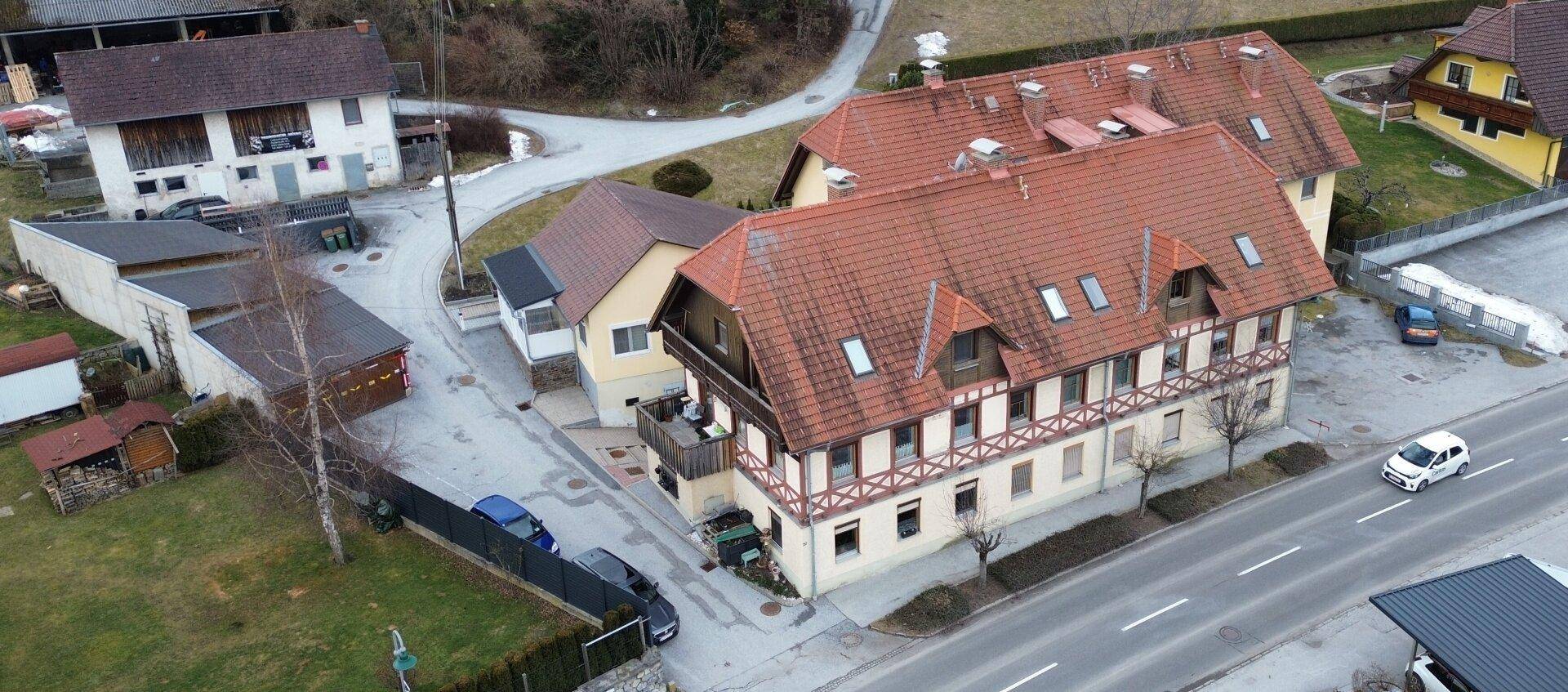 Blick von oben auf ein Wohngebäude mit mehreren Dächern, umgeben von Grünflächen und einer Straße.