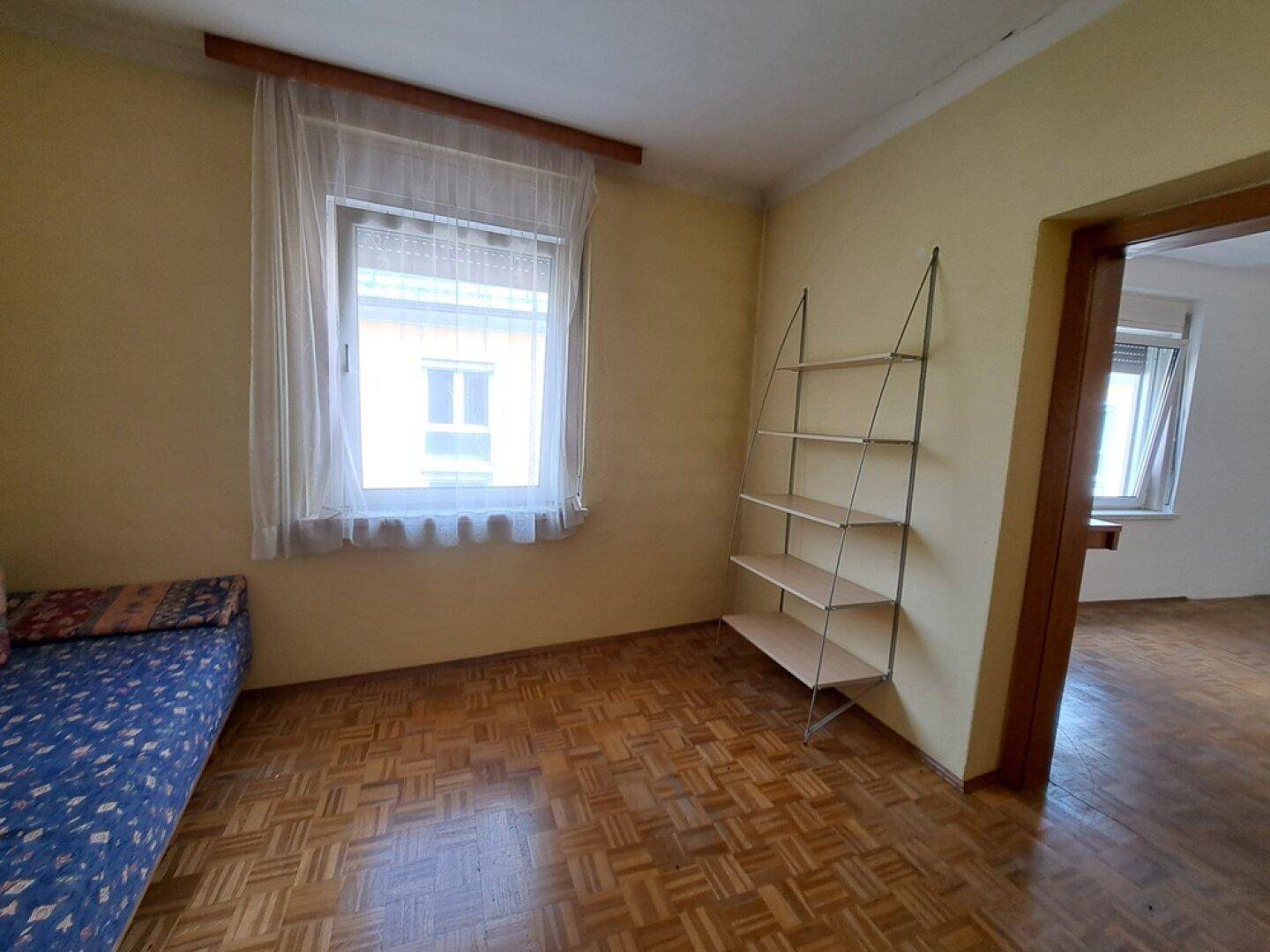 Helles Schlafzimmer mit Parkettboden, Fenster und einem einfachen Regal an der Wand.