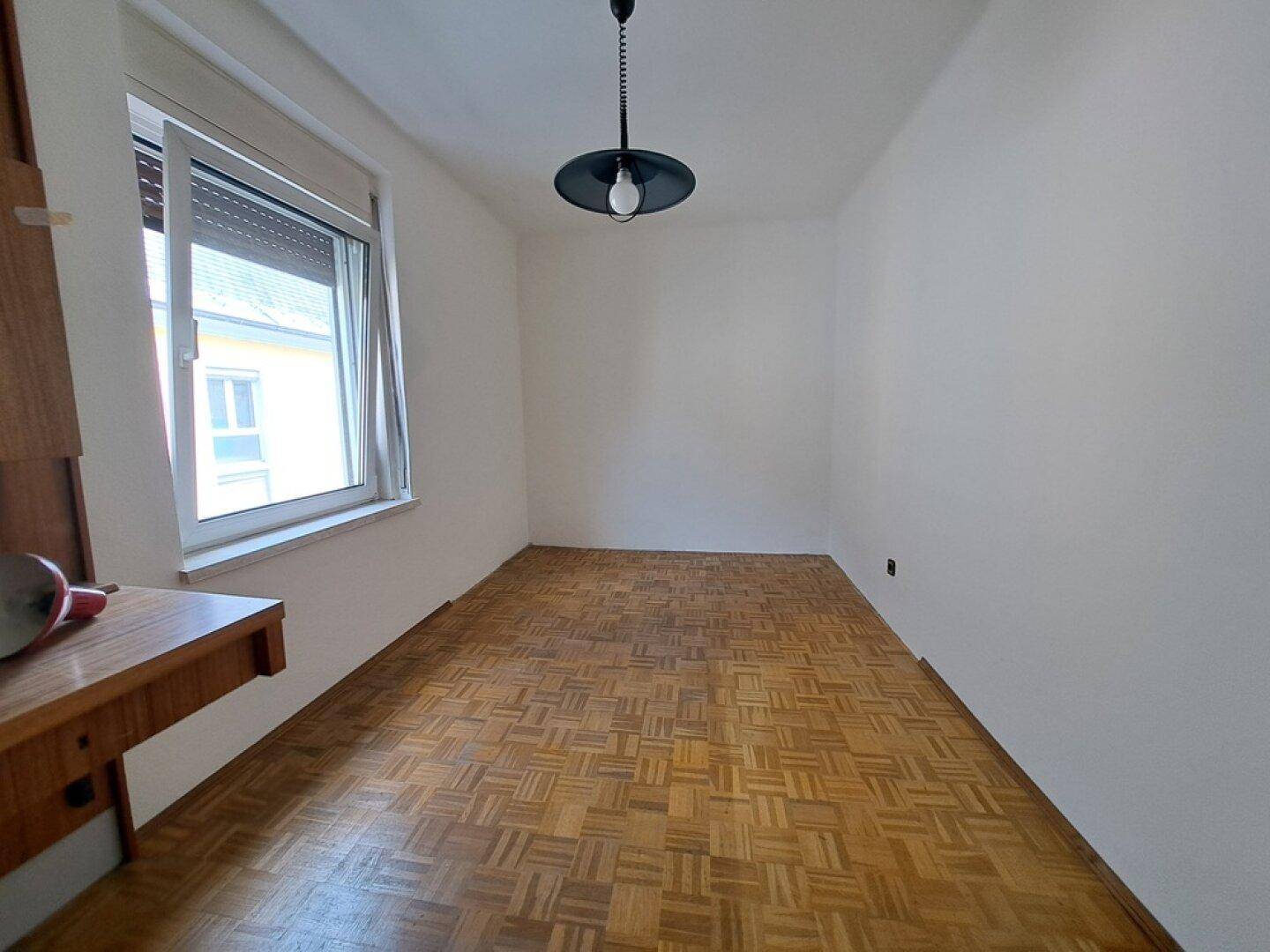 Leeres Schlafzimmer mit Parkettboden, weiß gestrichenen Wänden und einem Fenster.