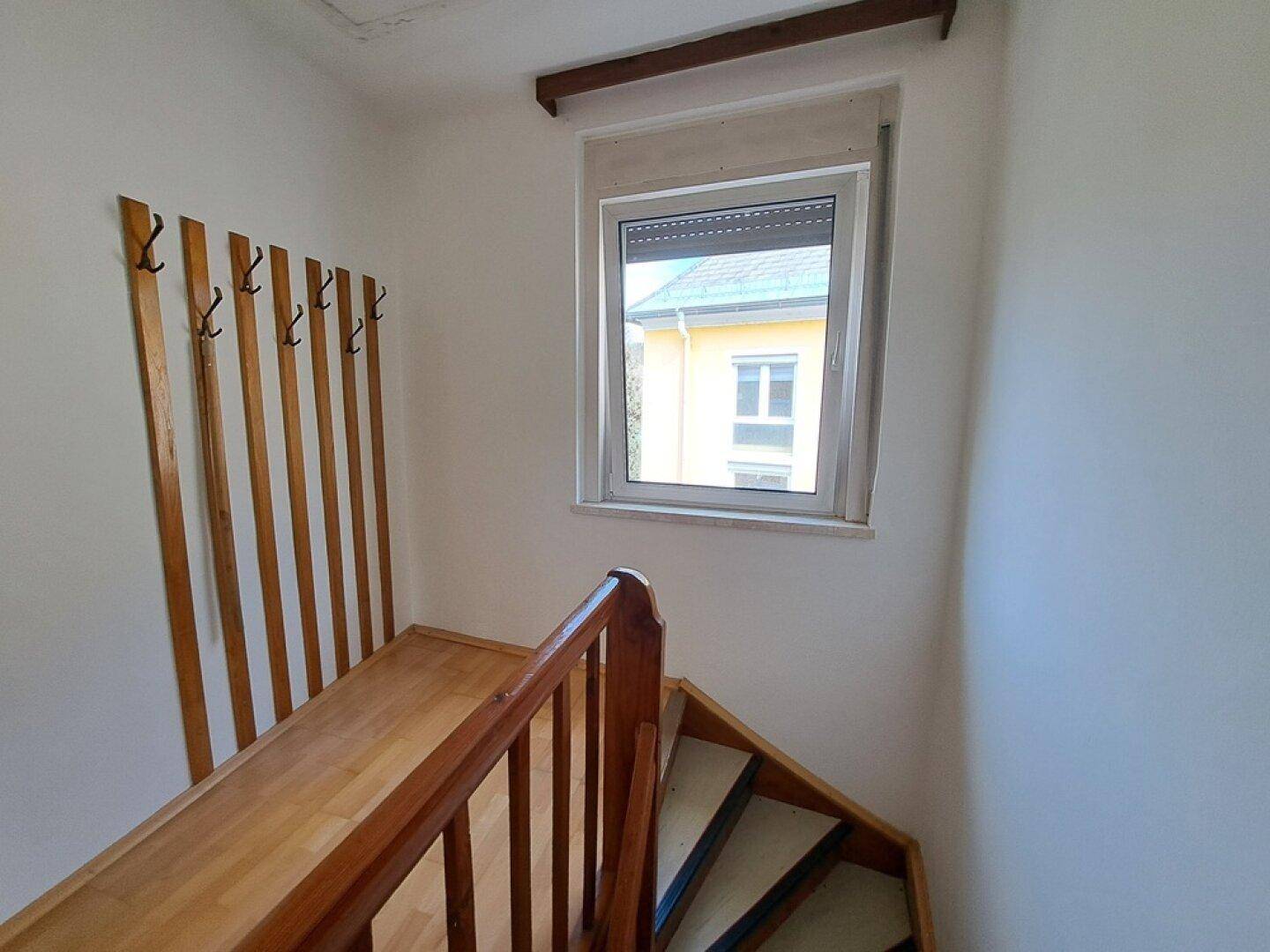 Treppenaufgang mit Holzgeländer, Garderobe und Fenster, das Tageslicht hereinlässt.