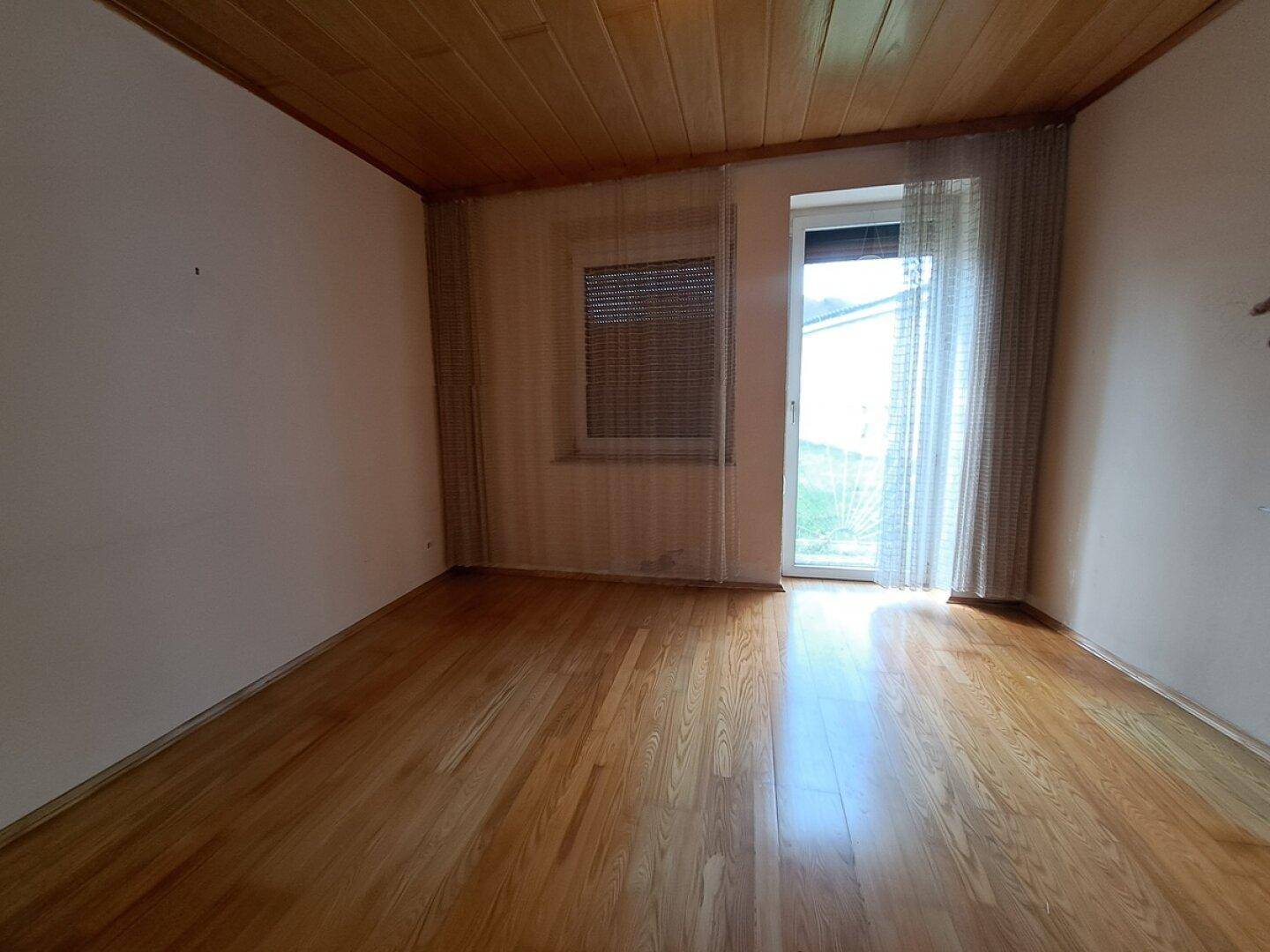 Leeres Zimmer mit hellem Parkettboden, Holzdecke und Zugang zum Garten durch eine Fenstertür.