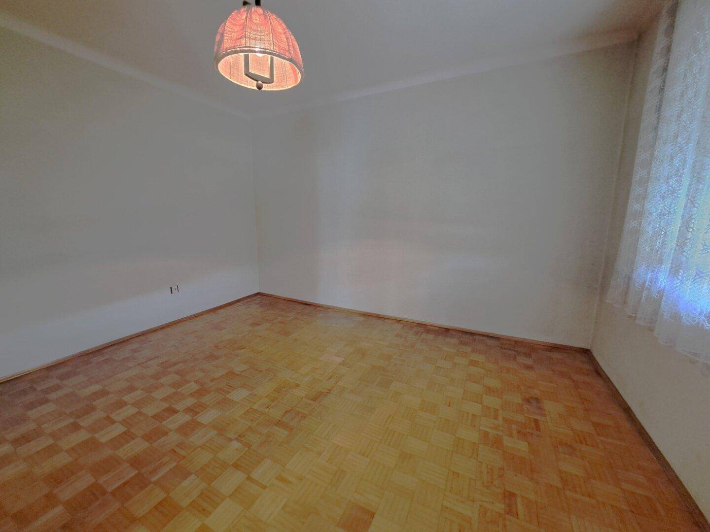 Schlafzimmer mit Parkettboden, weißen Wänden und einem Fenster mit Vorhängen.
