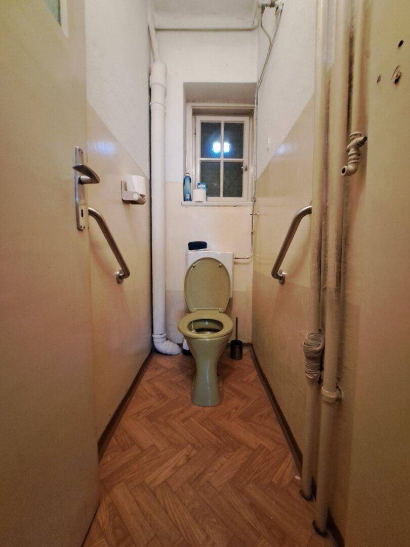 Schmales WC mit Fenster, Haltegriffen und einem WC in Pastellfarbe.