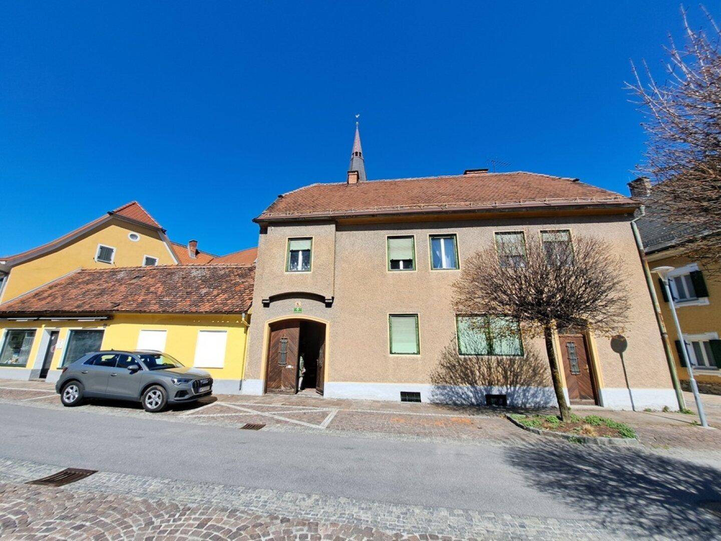 Fassade eines alten Gebäudes mit einem geparkten Auto davor und einem Kirchturm im Hintergrund.