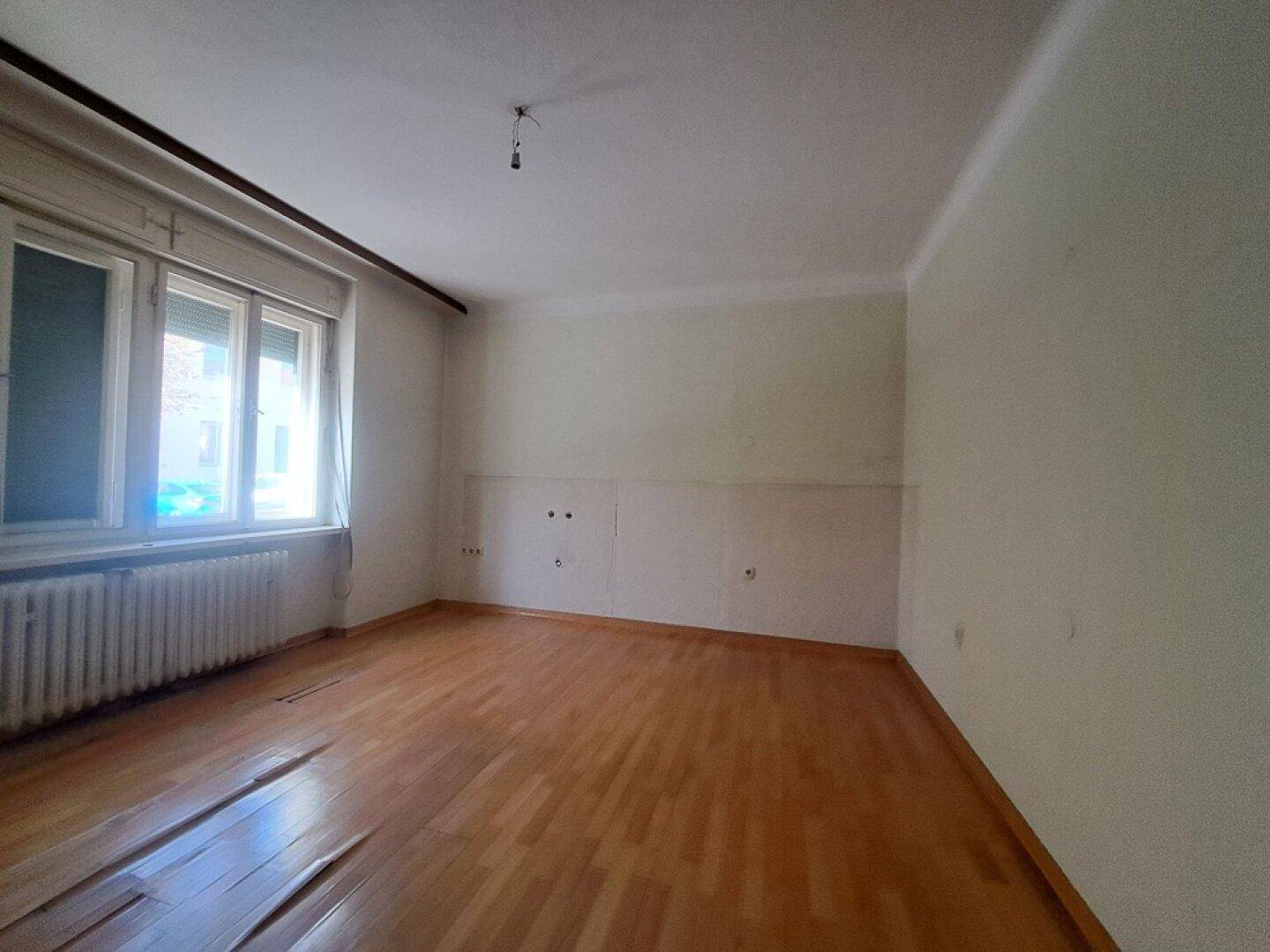 Heller, leerer Raum mit Holzfußboden, großen Fenstern und einem Heizkörper unter dem Fenster.