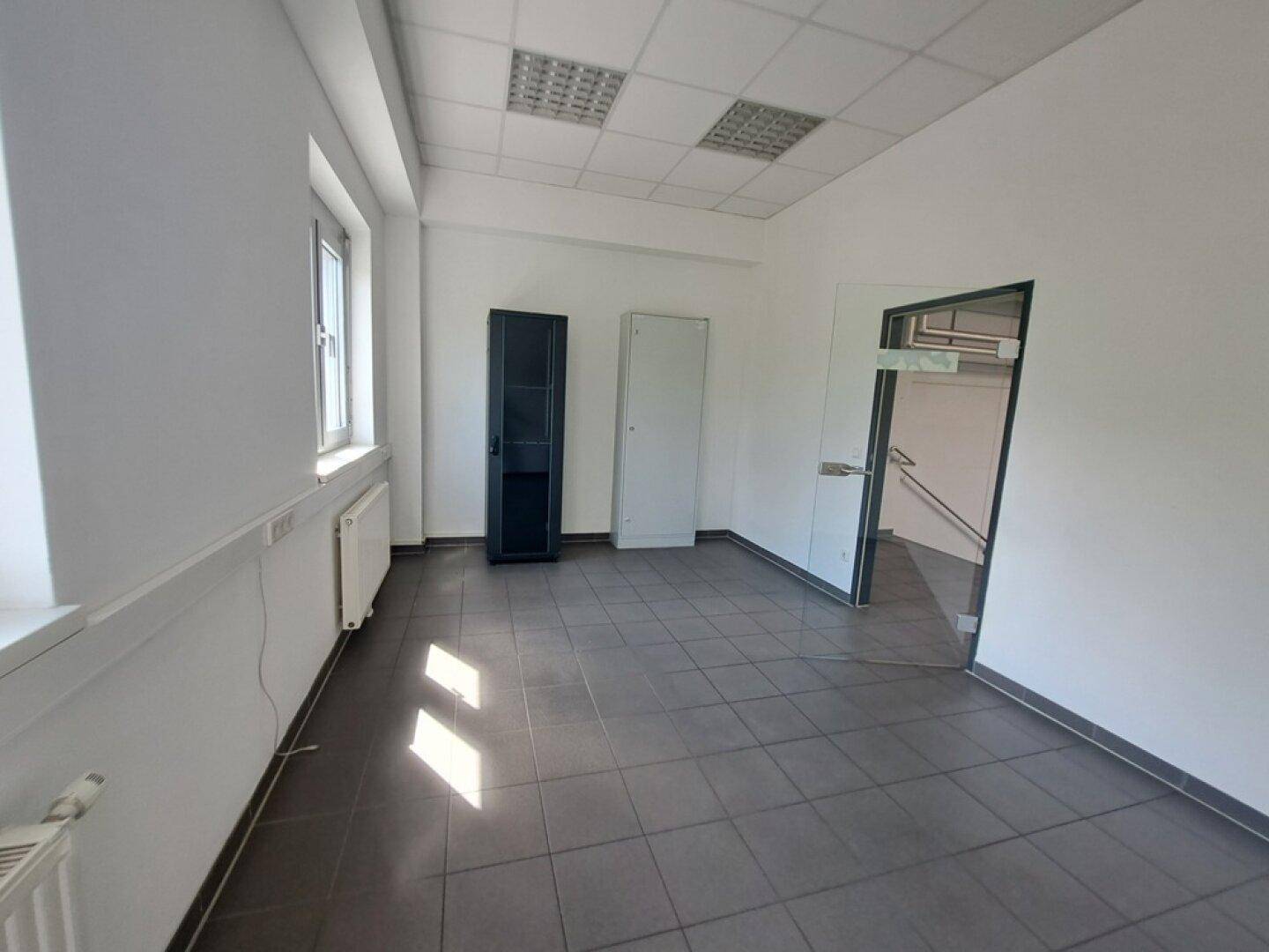 Leeres Büro mit dunklen Fliesen, weißen Wänden, einem Fenster und zwei Schränken.