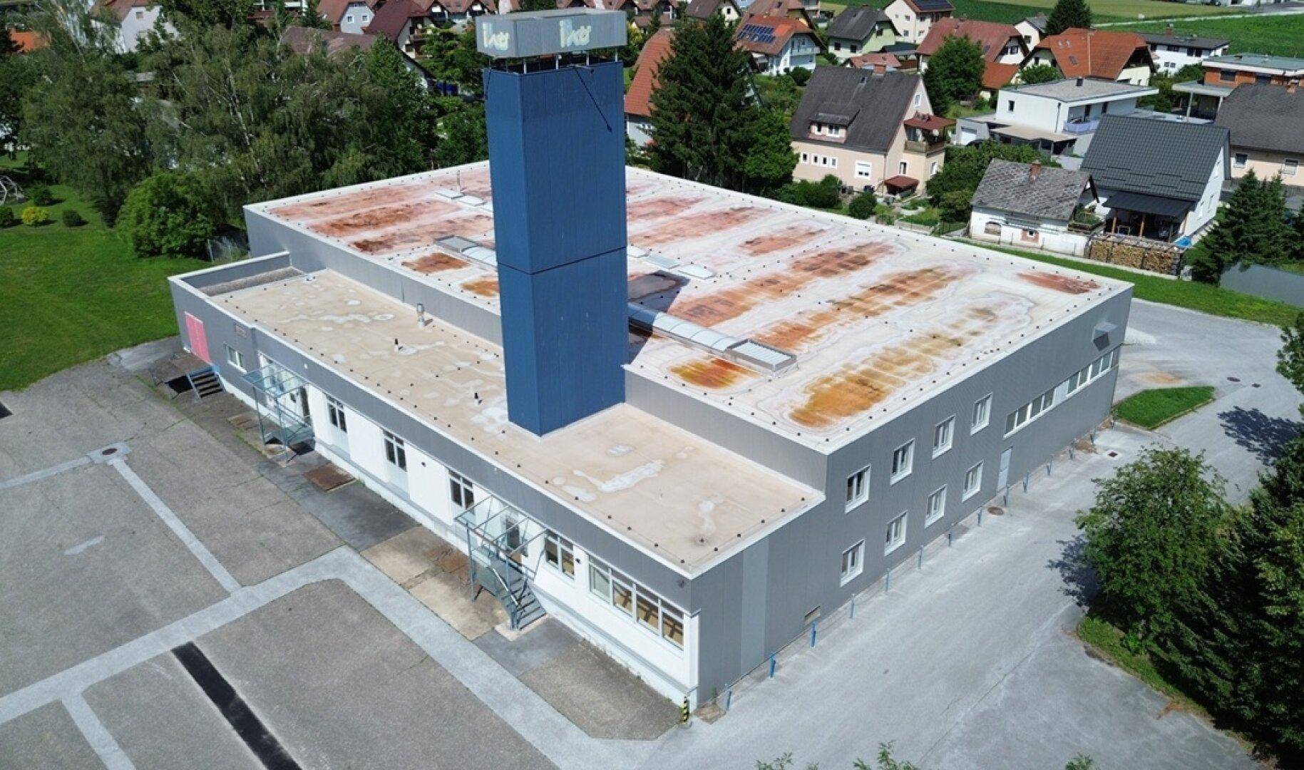 Luftaufnahme eines Gewerbegebäudes mit Flachdach, hohem blauen Schornstein und umliegender Wohnbebauung.