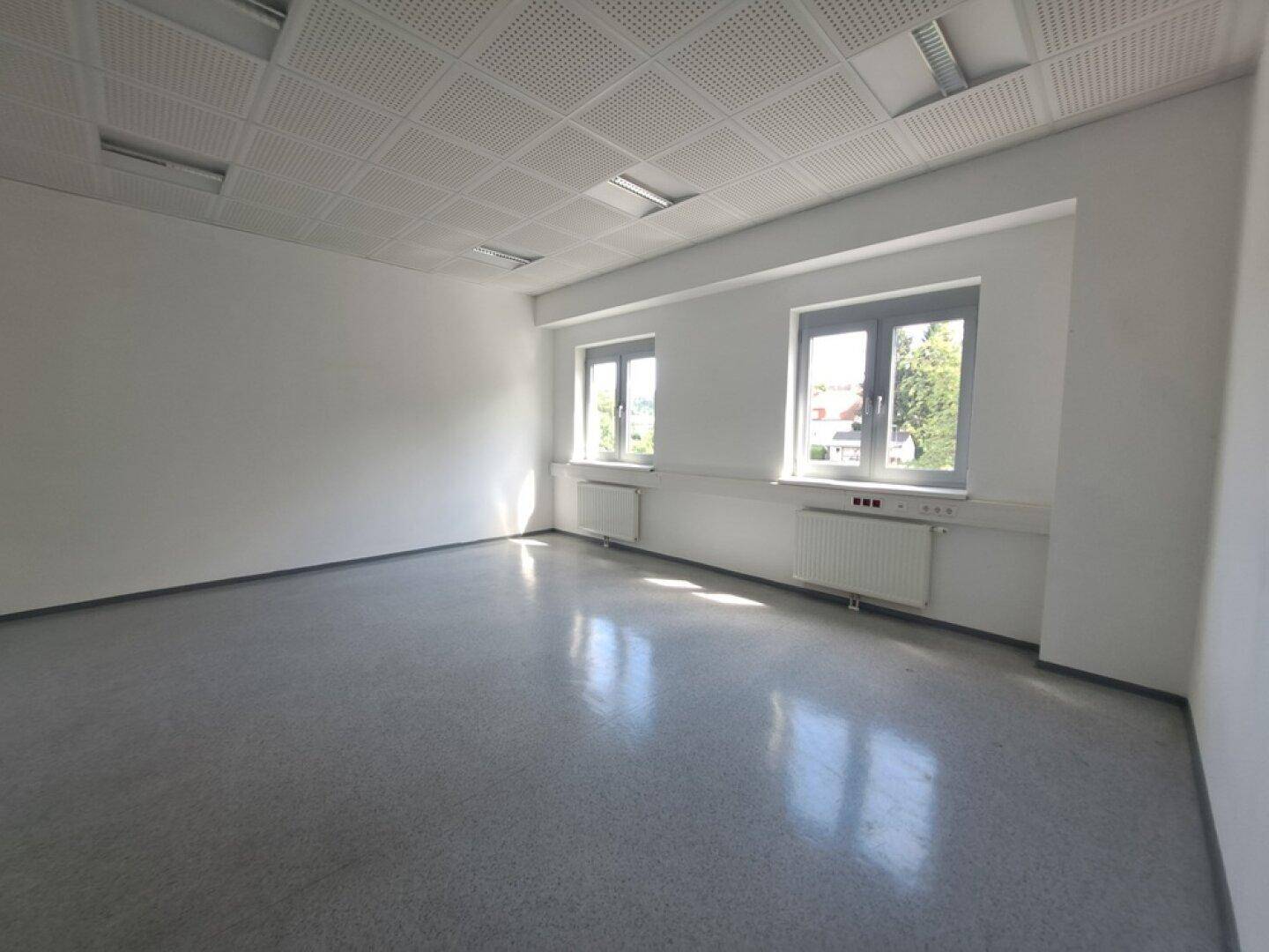 Helles, leeres Büro mit hellem Boden, weißen Wänden, zwei Fenstern und Heizkörpern.