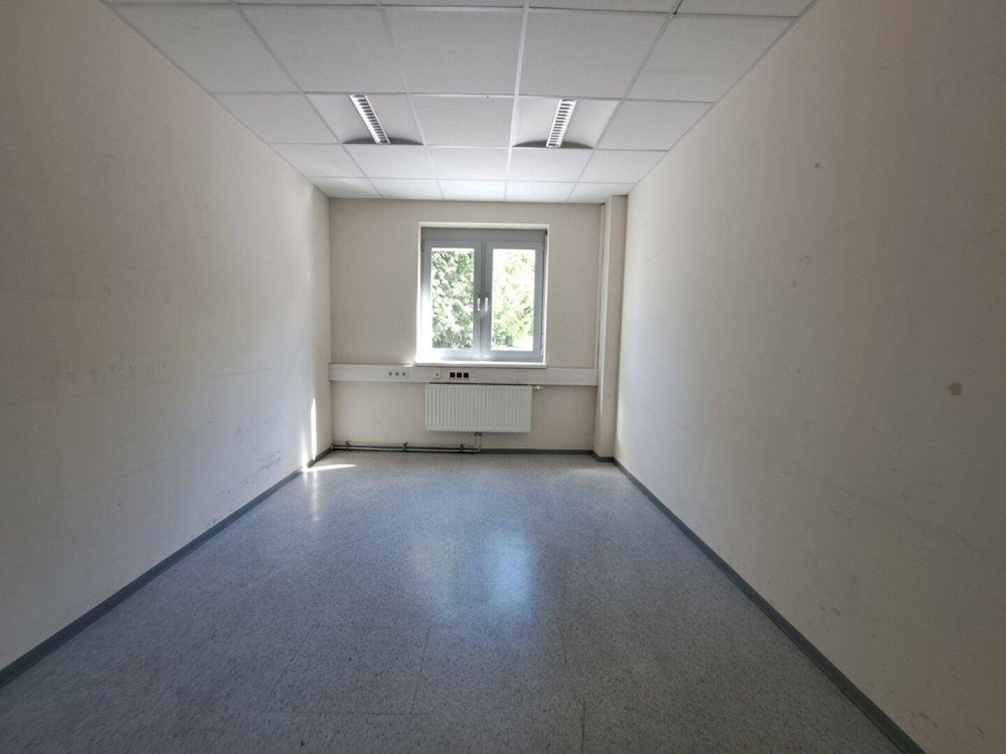 Schmaler, leerer Büroraum mit hellem Boden, weißen Wänden, einem Fenster und Heizkörper.