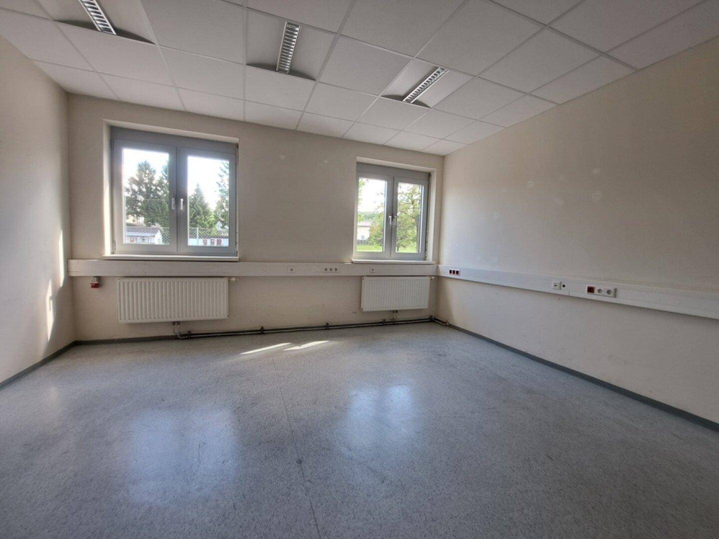 Helles, leeres Büro mit hellem Boden, weißen Wänden, zwei Fenstern und Heizkörpern.