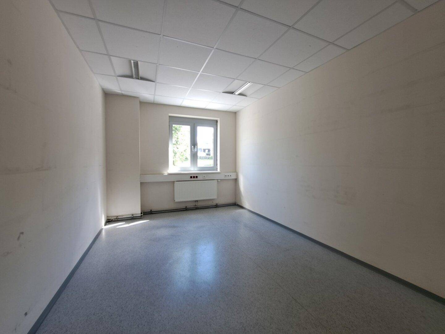 Leerer Büroraum mit hellem Boden, weißen Wänden und einem Fenster, das Tageslicht hereinlässt.